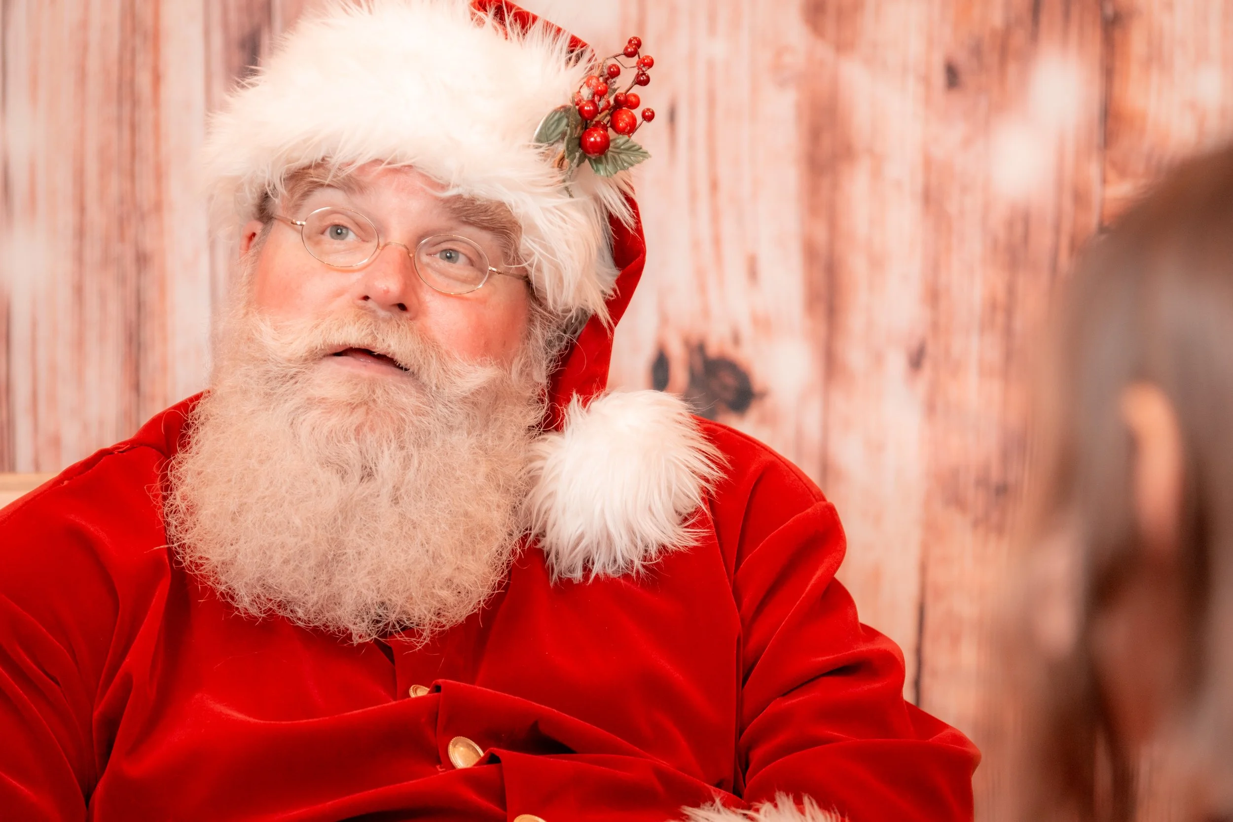 Santa Claus with glasses and a white beard, wearing a red velvet suit with white fur trim, and a Santa hat decorated with holly berries and leaves, sitting in front of a wooden background.
