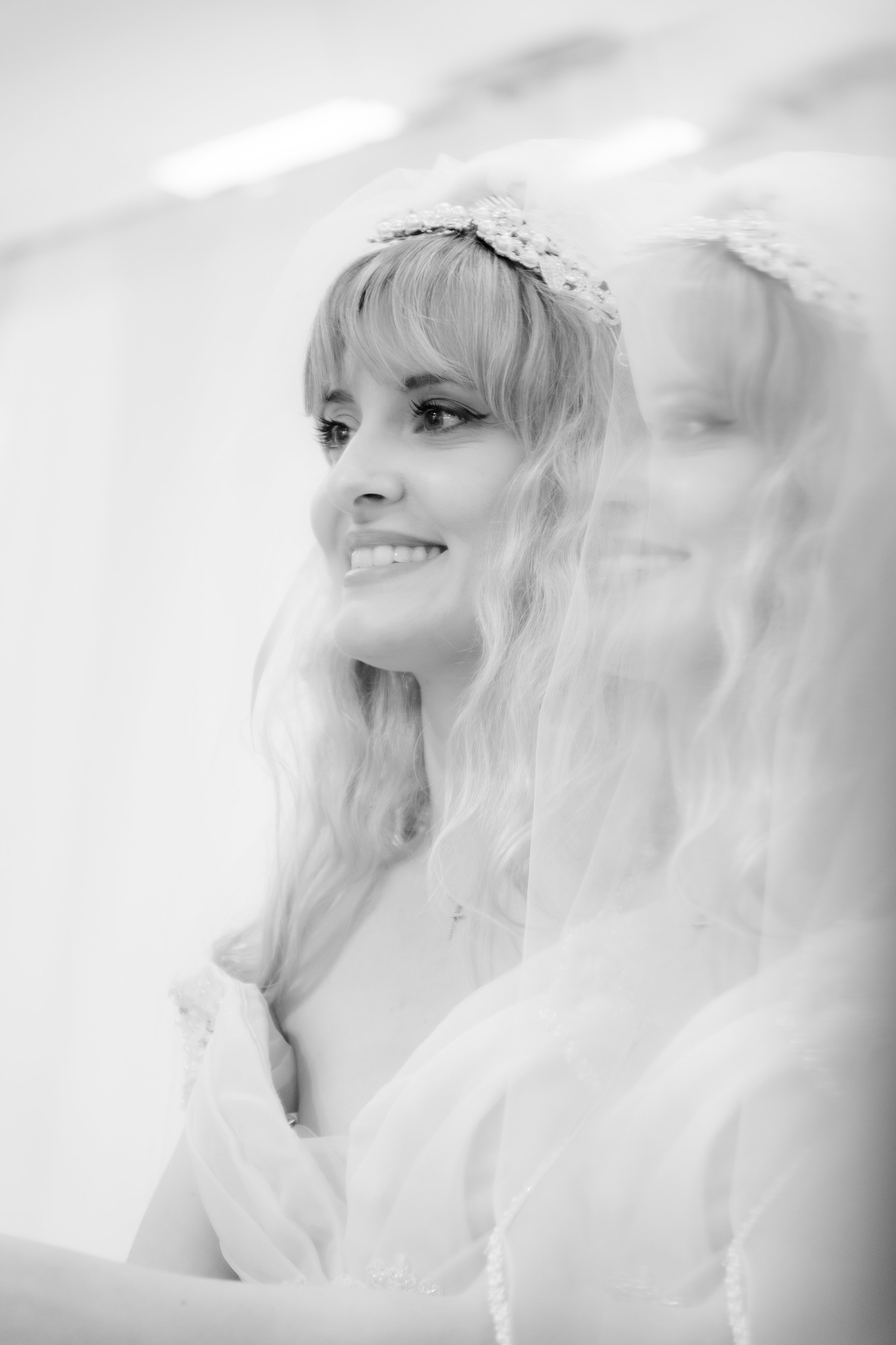 A smiling woman dressed as a bride, with wavy blonde hair and a floral headpiece, standing next to a reflection or another woman in a bridal veil.