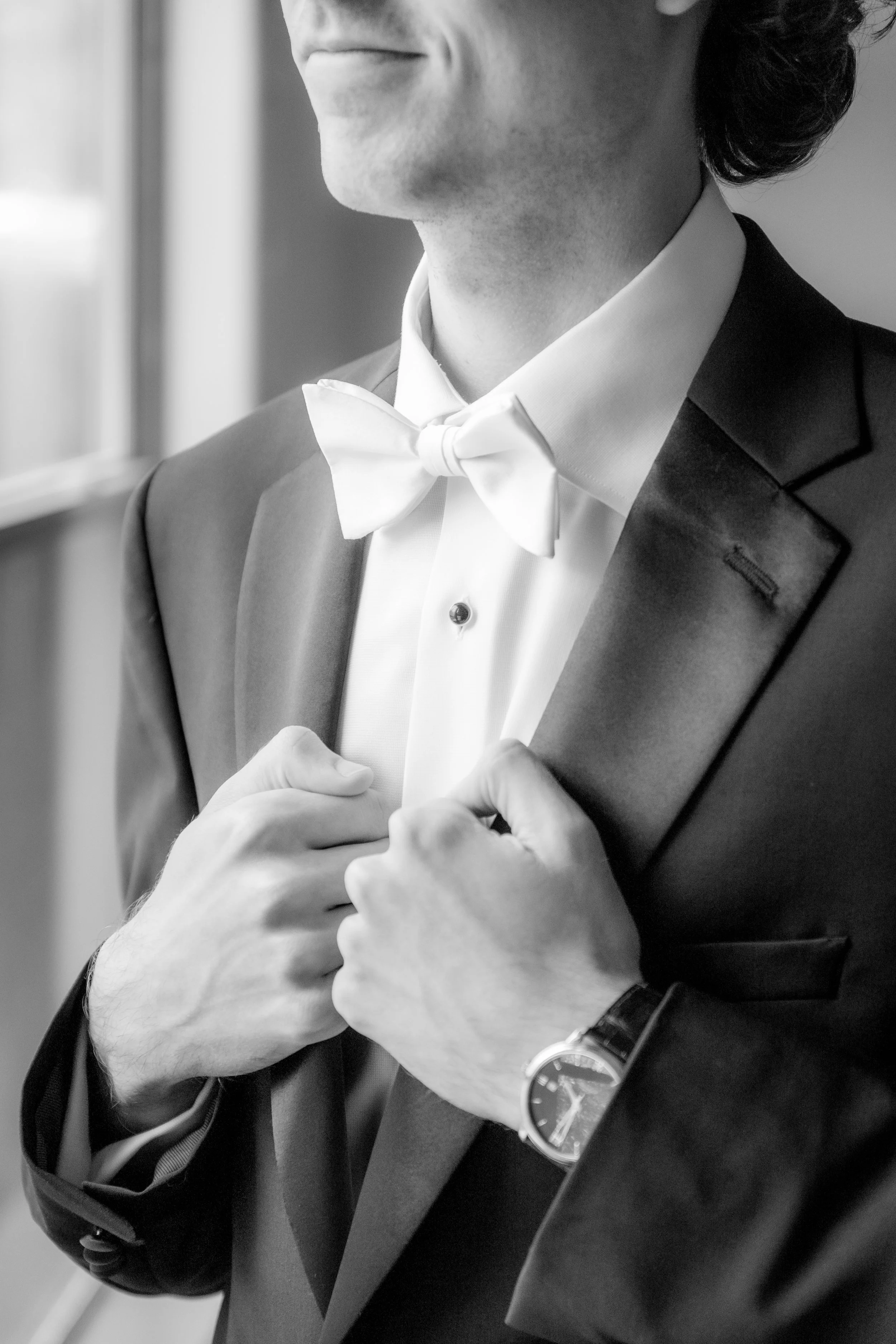A man in a tuxedo adjusting his suit jacket, wearing a white dress shirt, a bow tie, and a watch on his left wrist, with a window in the background.