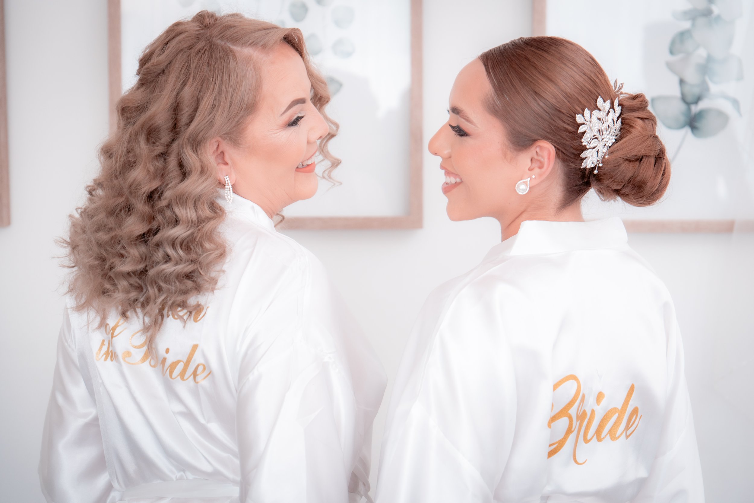 Two women dressed as brides smiling at each other, wearing white satin robes with 'Bride' embroidered in gold on the back, standing in a bright, decorated room.