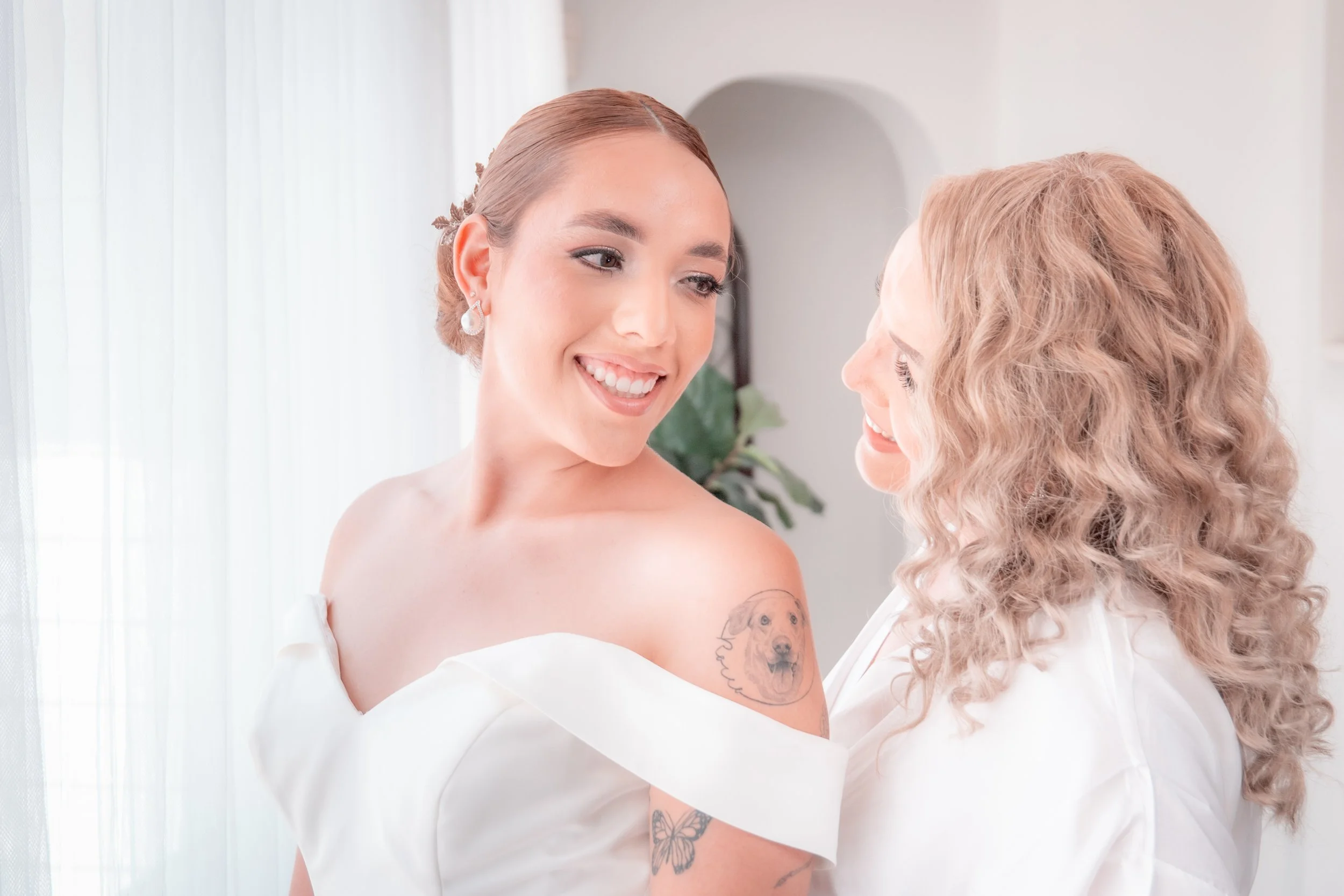 Two women dressed in white, smiling at each other, standing close inside a bright room with sheer curtains and a potted plant in the background.