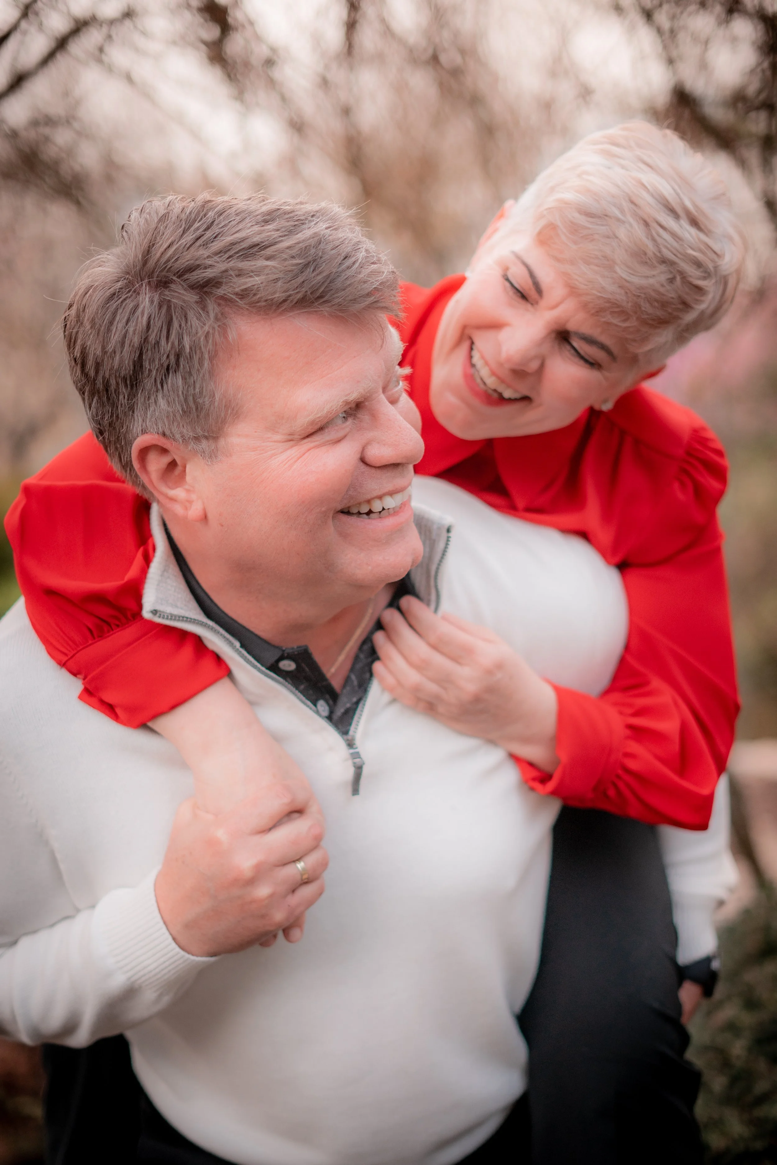 A senior man giving a piggyback ride to a smiling woman outdoors, both appearing joyful with trees in the background.