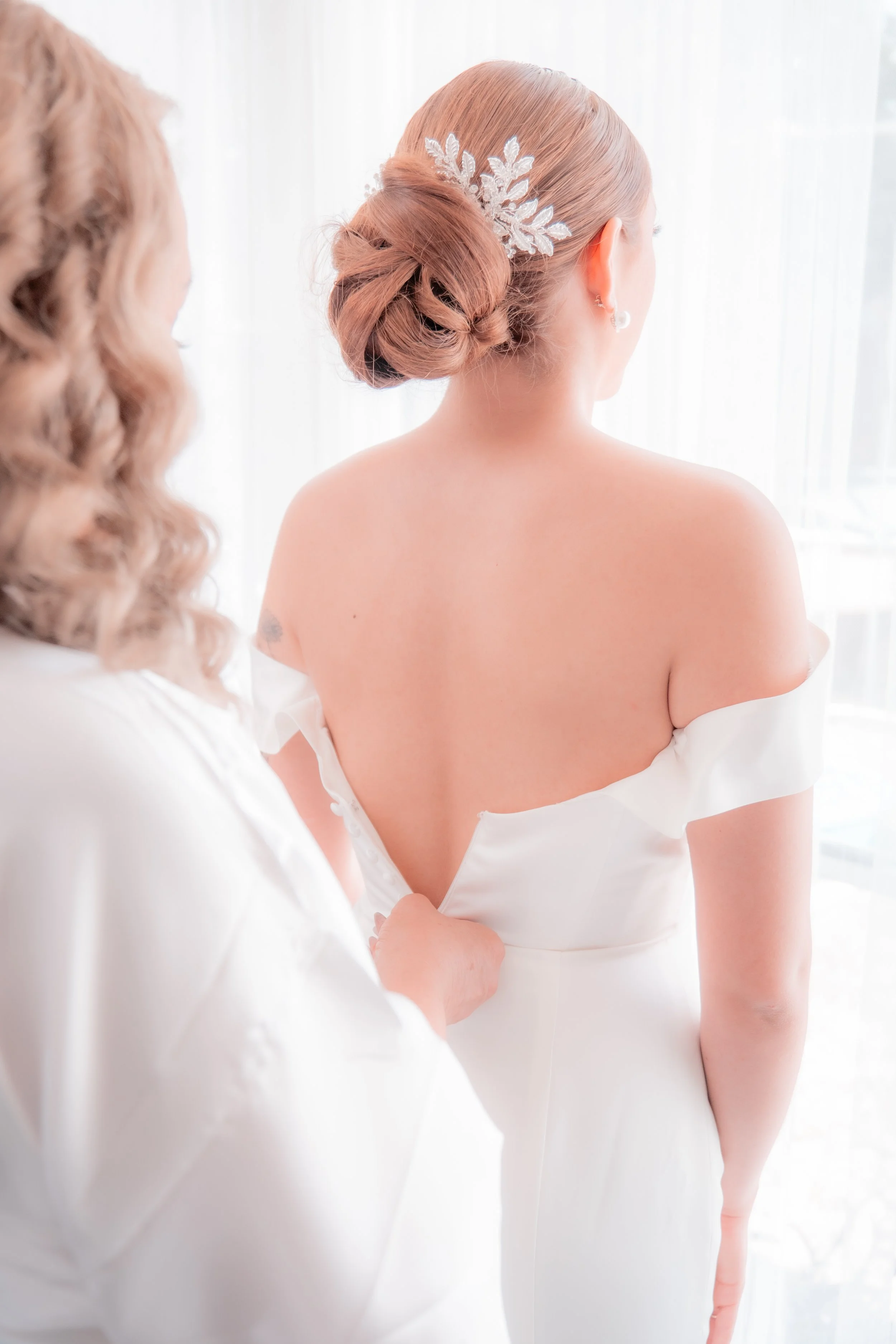 Bride with styled hair adding final touches to her wedding dress, standing near a window with sunlight.