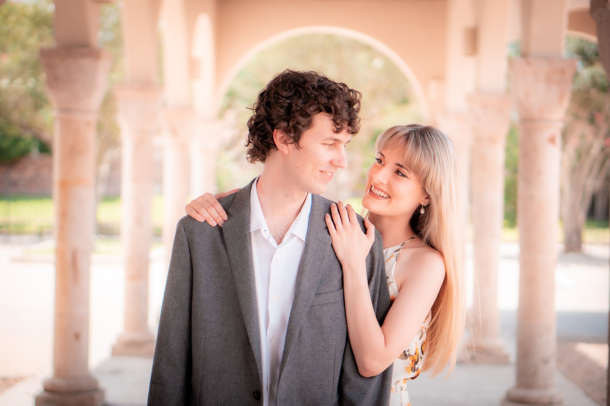 A young man and woman stand close together outdoors, smiling and looking at each other, with the woman resting her hand on the man's shoulder. They are in front of a background with stone columns and arches, with trees and sunlight visible.