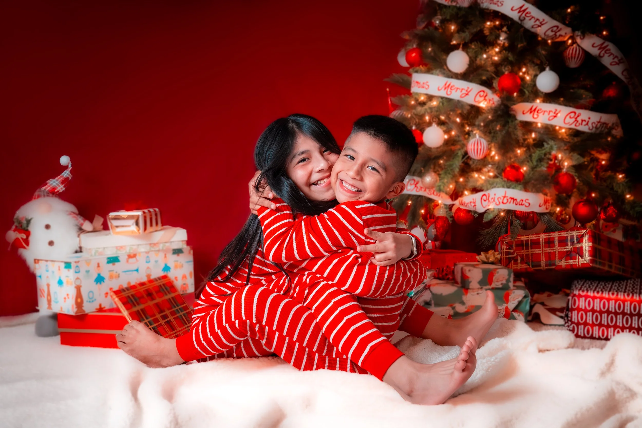 Two children, a boy and a girl, dressed in red and white striped pajamas, hugging and smiling in front of a decorated Christmas tree with presents underneath and a red wall in the background