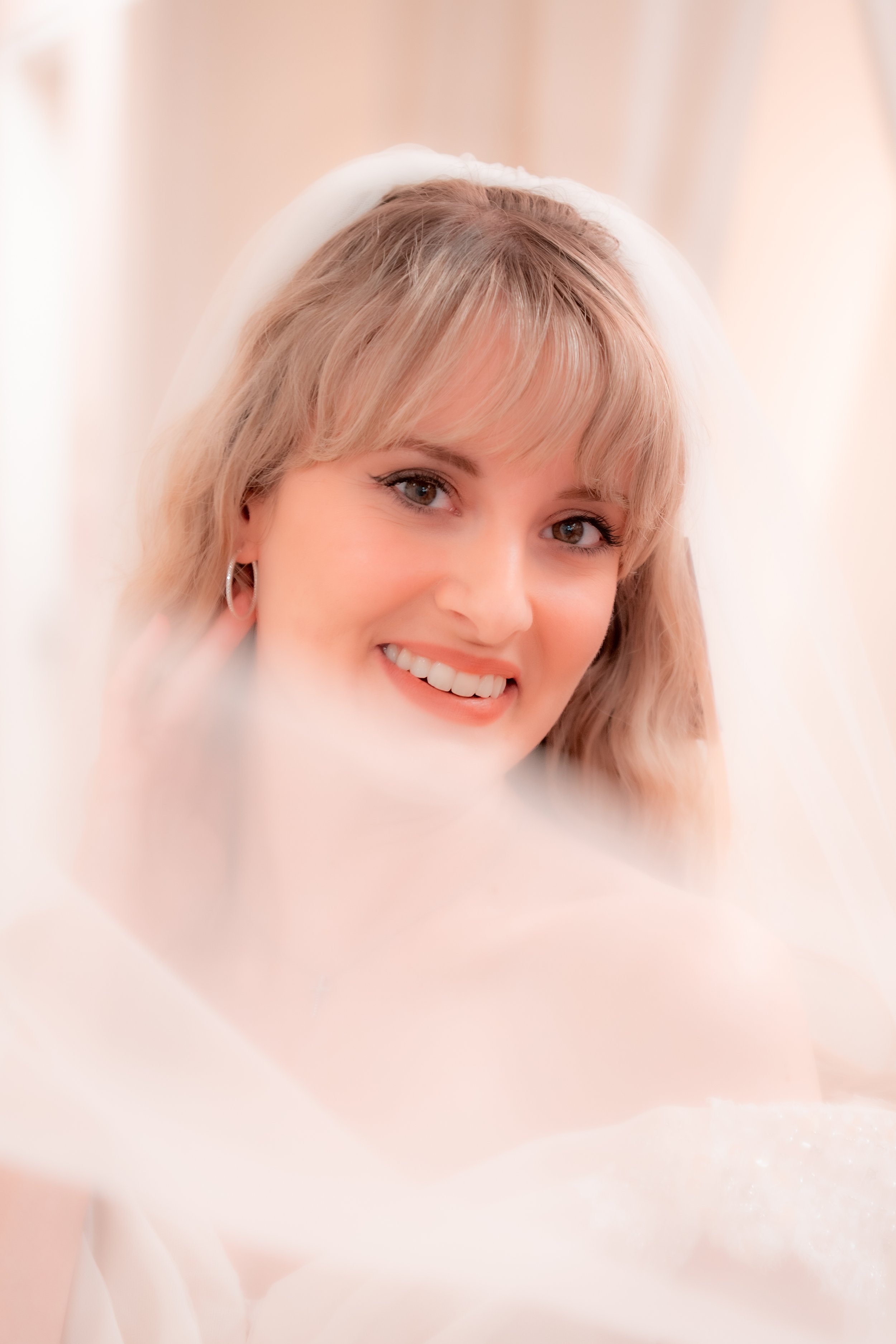 A smiling woman with blonde hair, wearing a bridal veil, in a soft, warm setting.