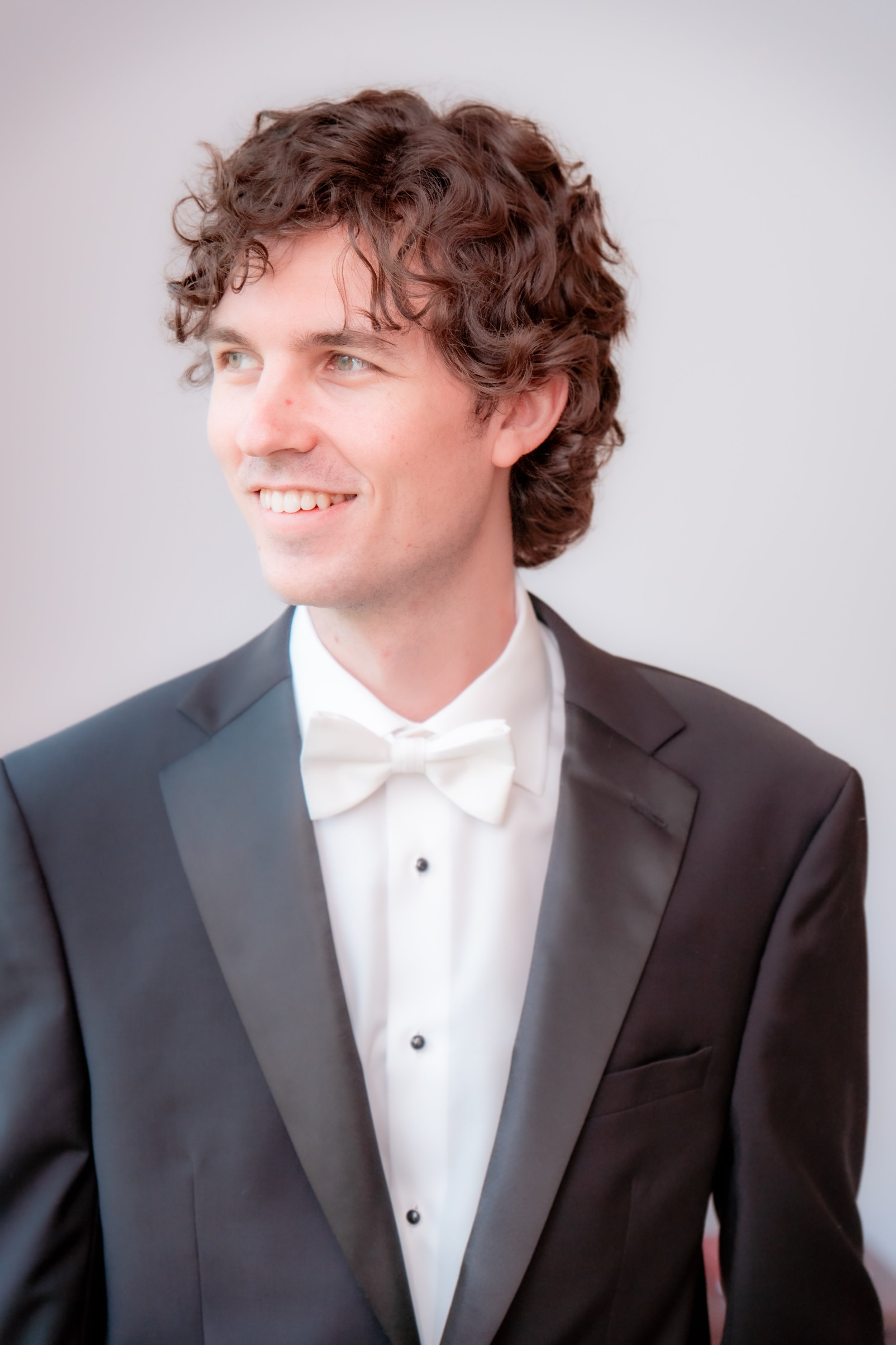 A young man with curly brown hair, wearing a black tuxedo with a white dress shirt and a white bow tie, smiling and looking to the side.