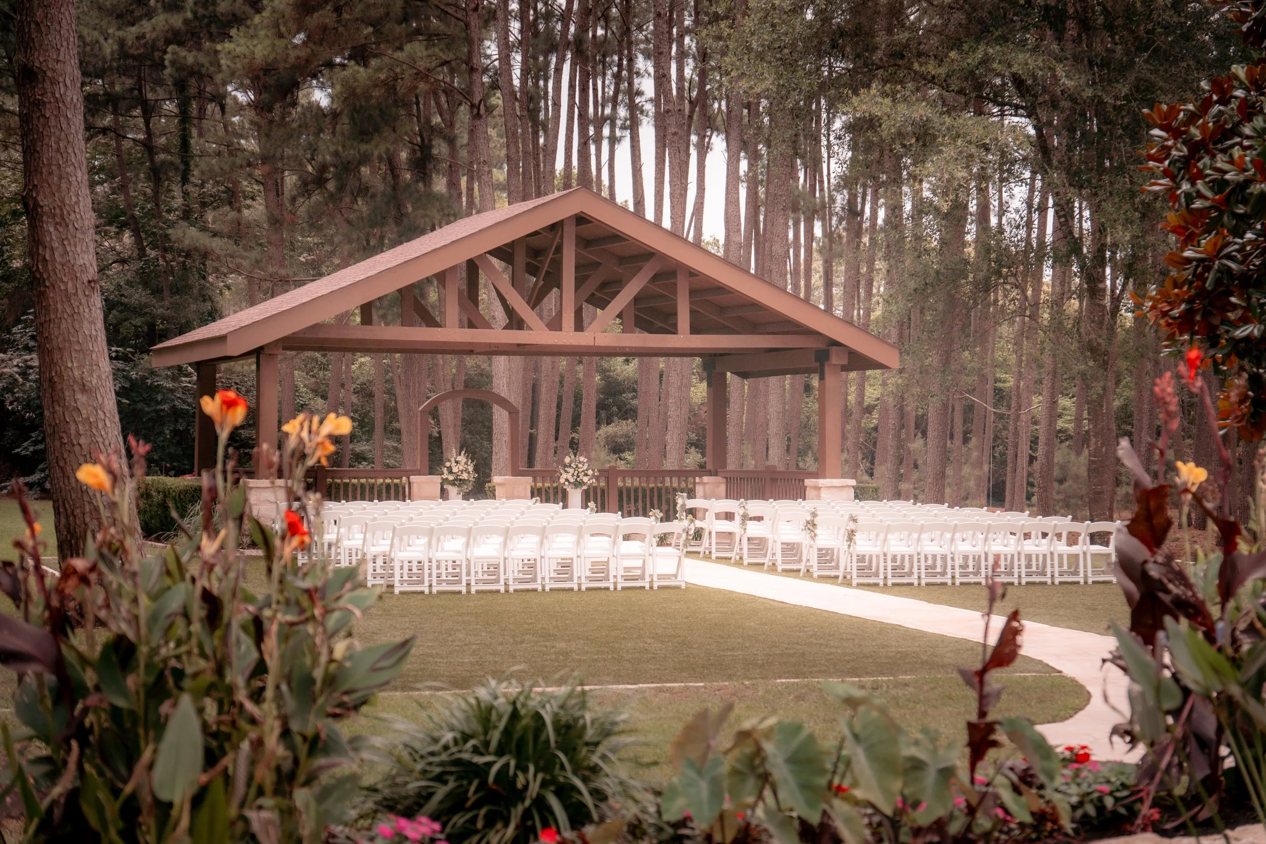 Outdoor wedding ceremony setup with white chairs arranged in rows on a lawn in front of a wooden stage under a pavilion, surrounded by trees and flowers.