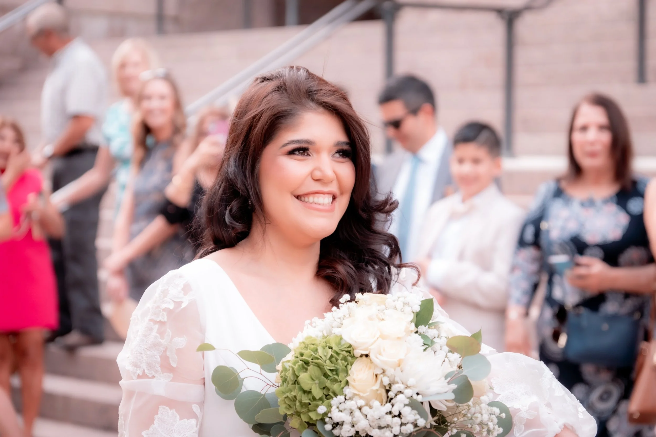 Smiling bride holding a bouquet of white flowers and greenery at a celebration with guests in the background.