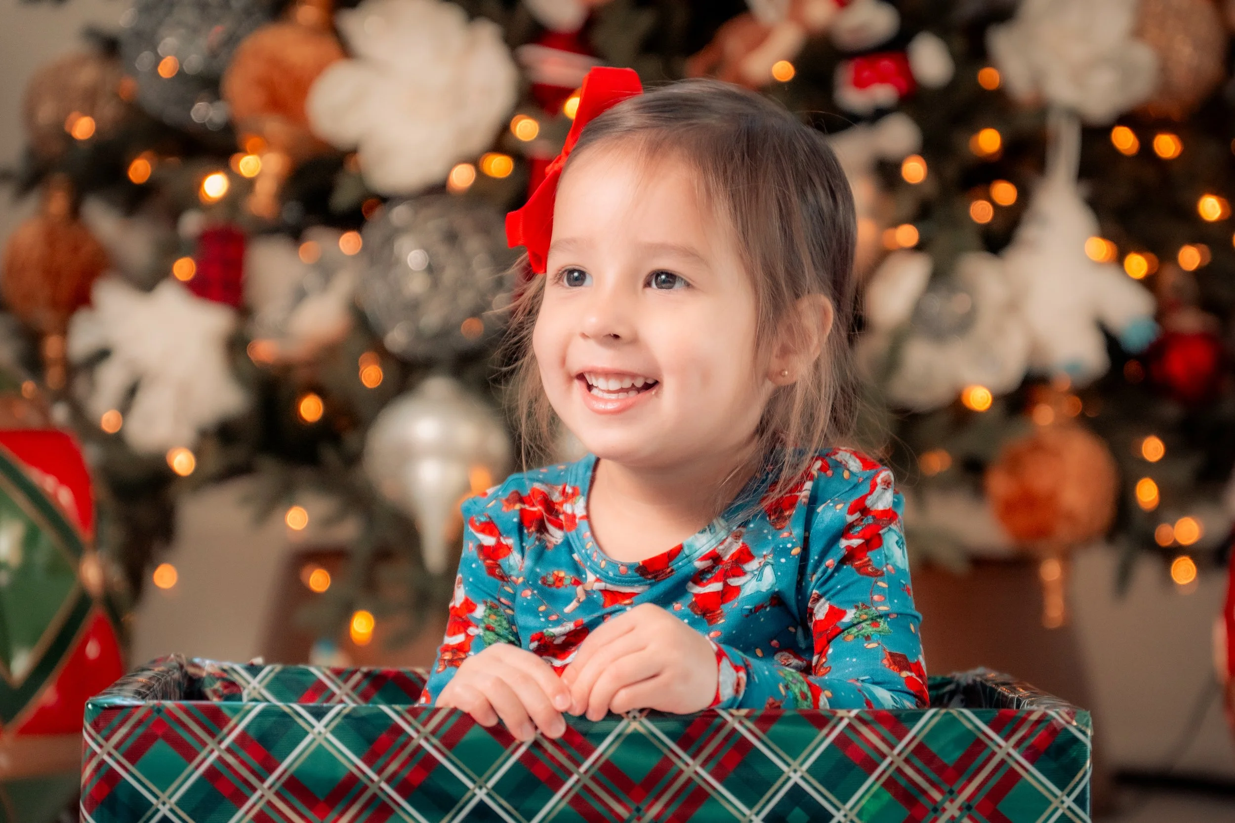 A smiling young girl wearing a Christmas-themed shirt and a red bow in her hair, sitting inside a holiday gift box in front of a decorated Christmas tree with ornaments and lights.