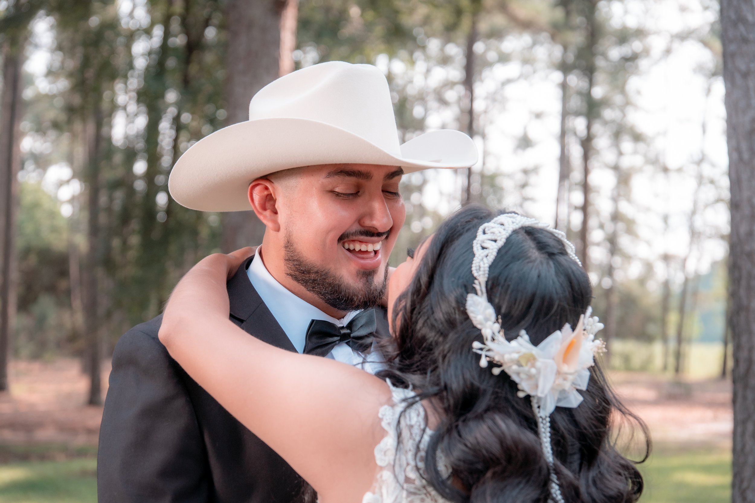 A couple in wedding attire embracing outdoors, with the man wearing a white cowboy hat and the woman wearing a floral headpiece, surrounded by trees.