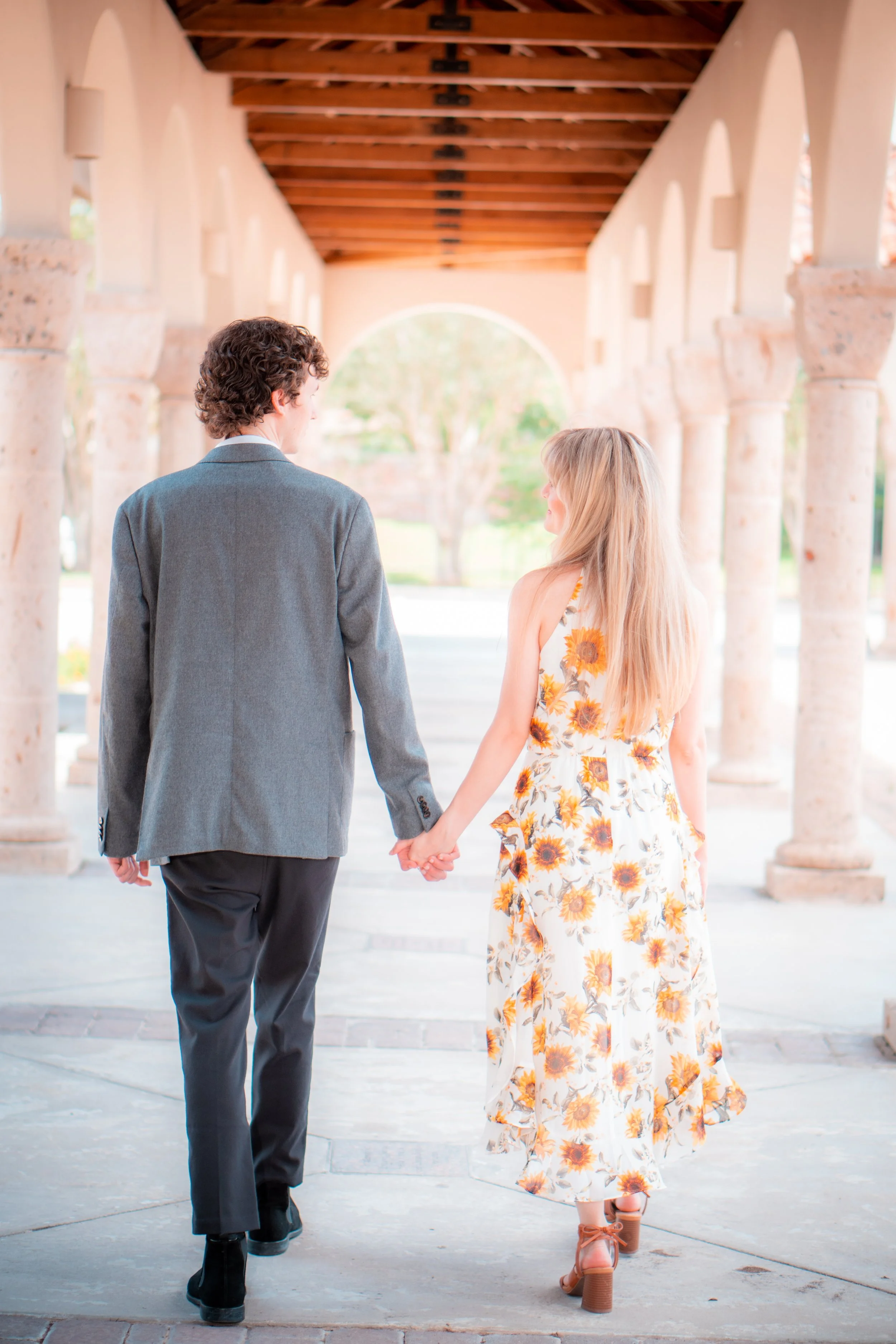A young couple, a man in a gray blazer and black pants, and a woman in a white sunflower dress and brown heels, holding hands as they walk under a stone archway in a park.