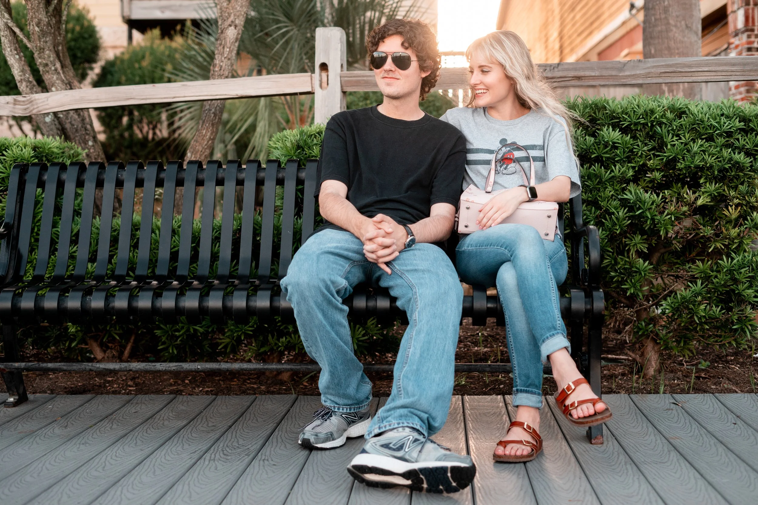 A young couple sitting together on a black metal park bench in a green outdoor area with trees and shrubs. The man wears sunglasses, a black t-shirt, jeans, and sneakers. The woman has blonde hair, a gray t-shirt, jeans, and sandals. She is smiling a
