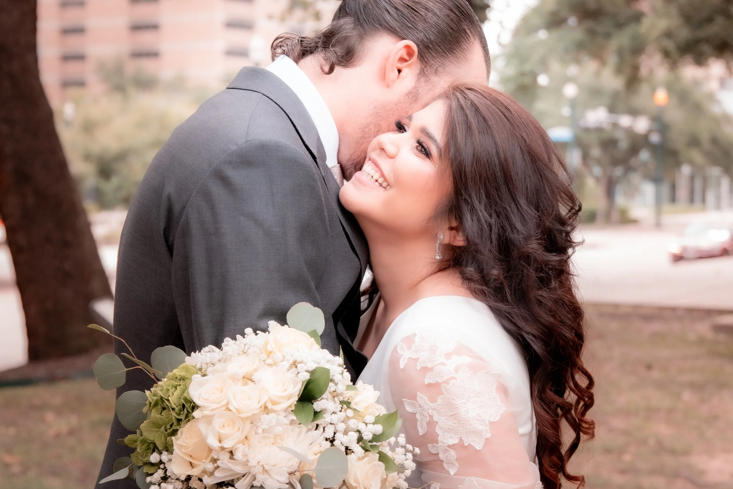 A bride and groom sharing a kiss outdoors, with the bride holding a bouquet of white roses and greenery.