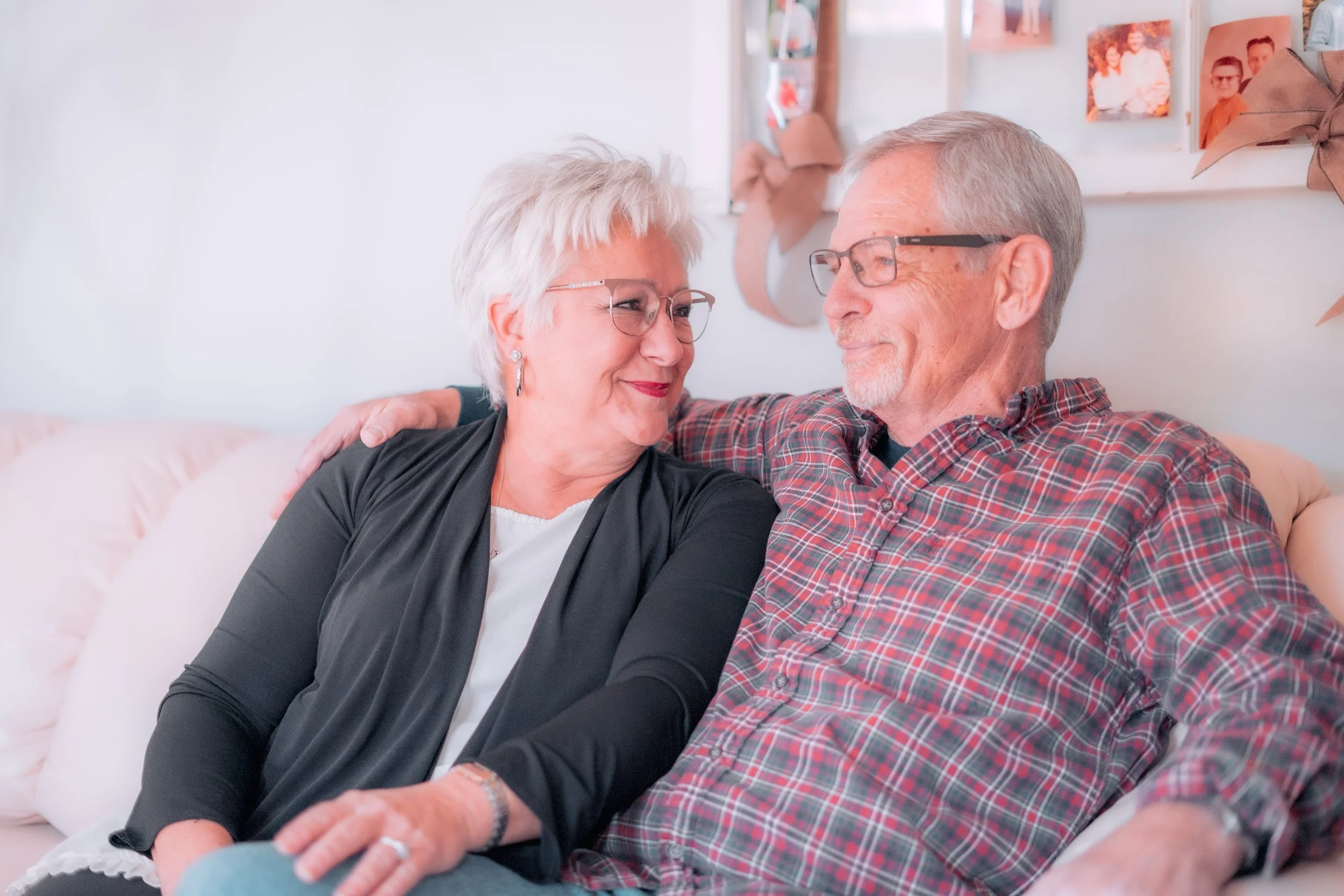 An elderly couple sitting close on a couch, smiling at each other, in a cozy, decorated room.