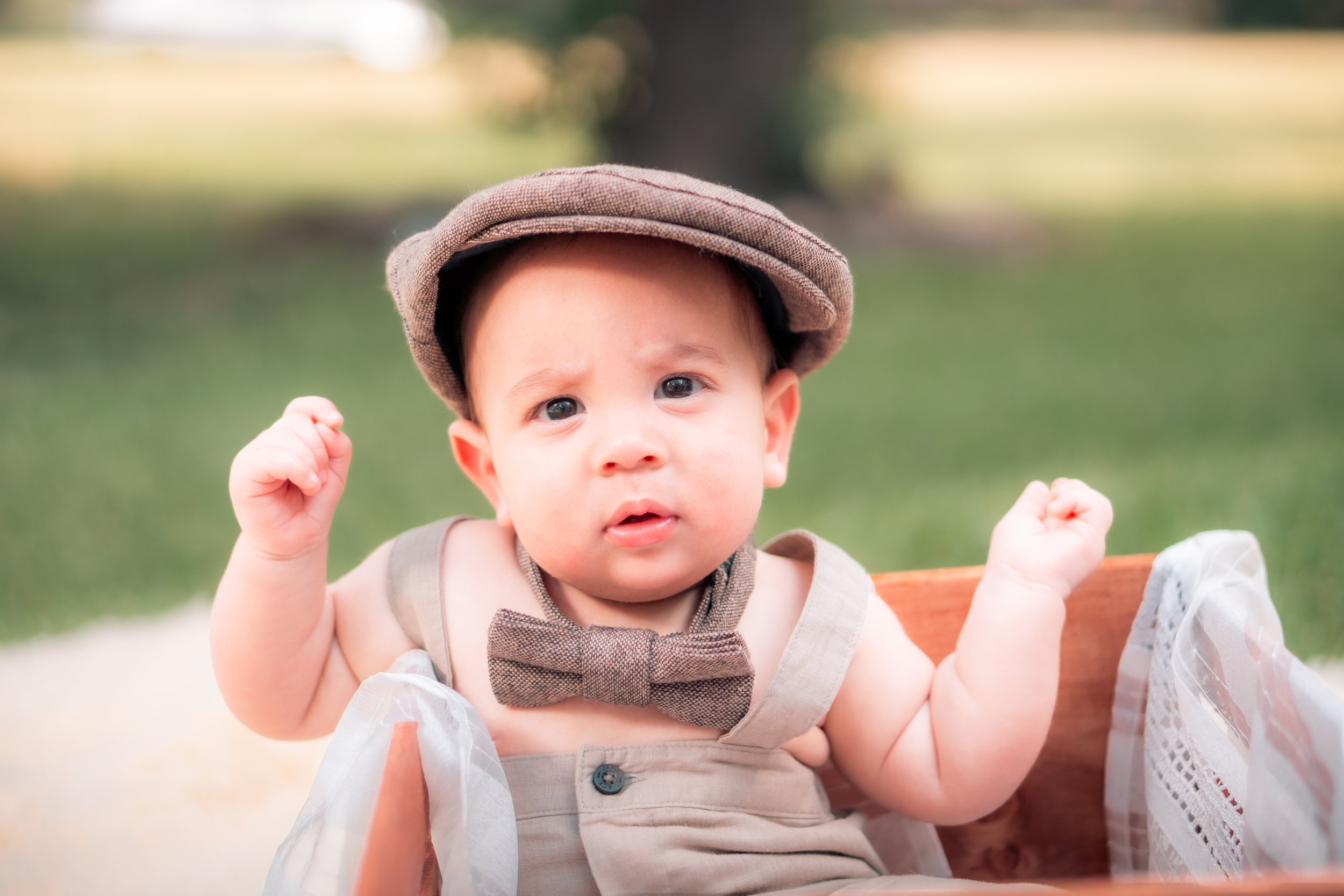 A baby dressed in vintage-style clothing, including a bow tie and matching hat, sitting in a basket outdoors with a blurred grassy background.