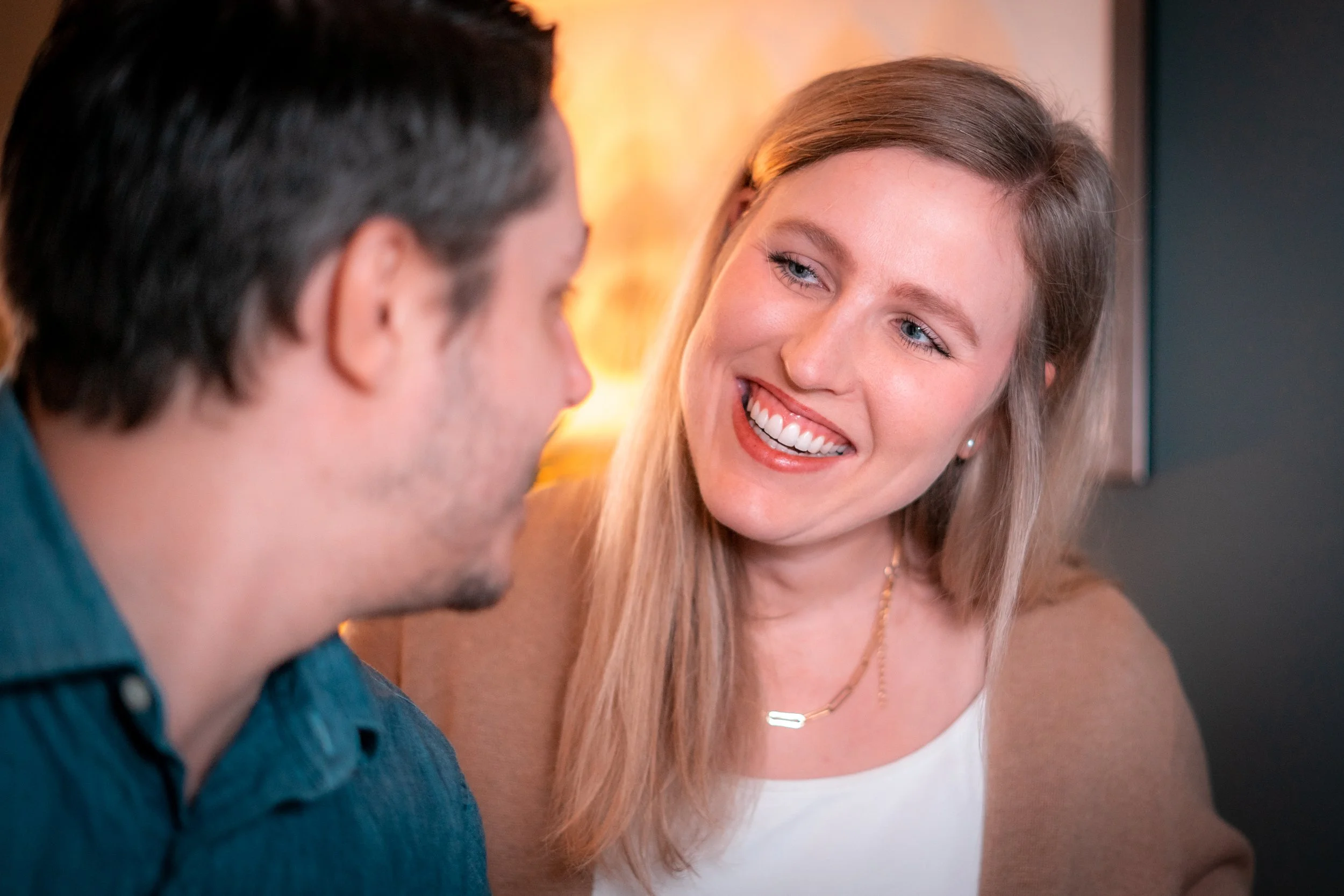 A woman smiling and talking to a man in a cozy indoor setting.