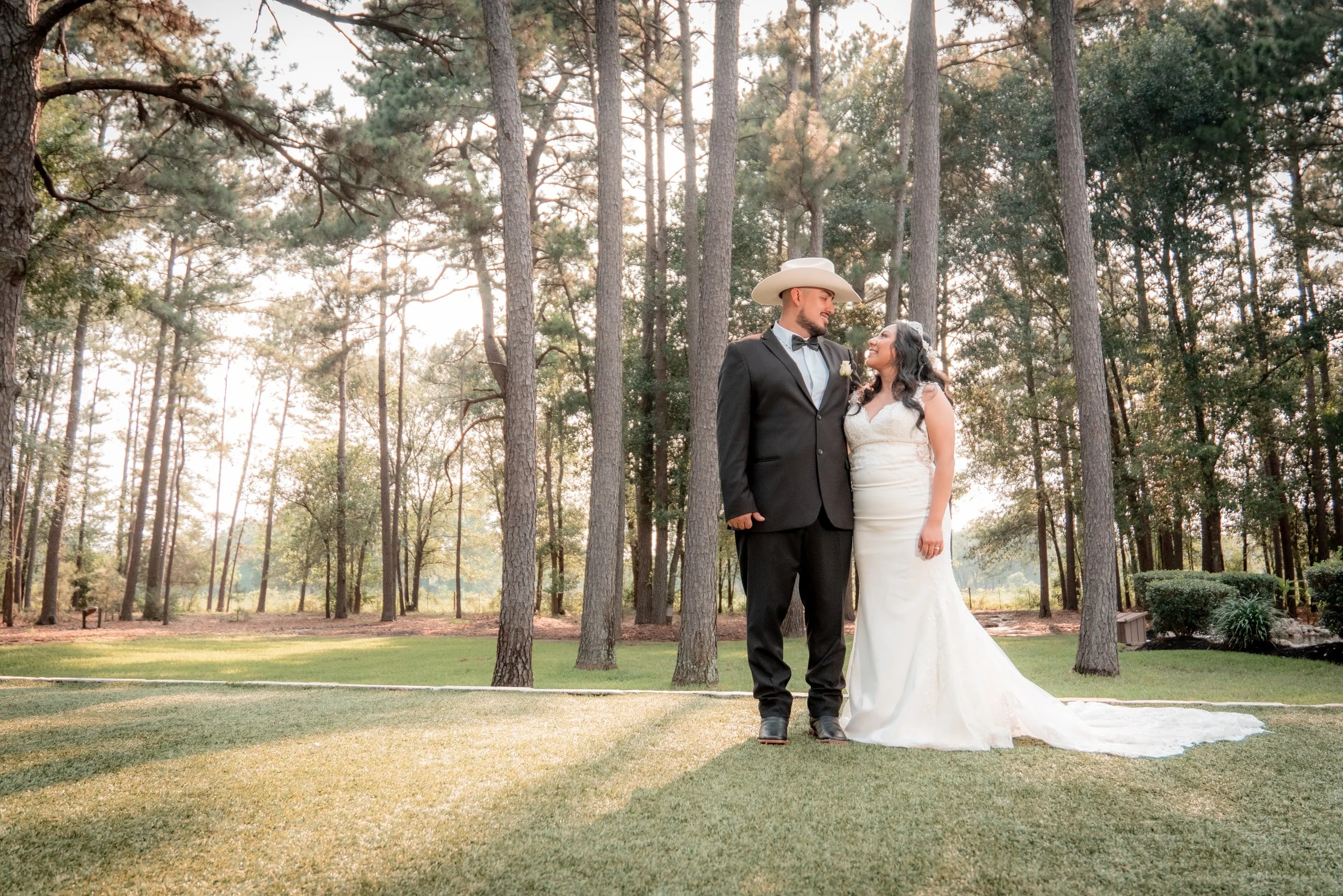 A newly married couple standing on a grassy area in a wooded park during sunset. The groom is wearing a black suit with a bow tie and a white cowboy hat, and the bride is in a white wedding dress. They are looking at each other and smiling.