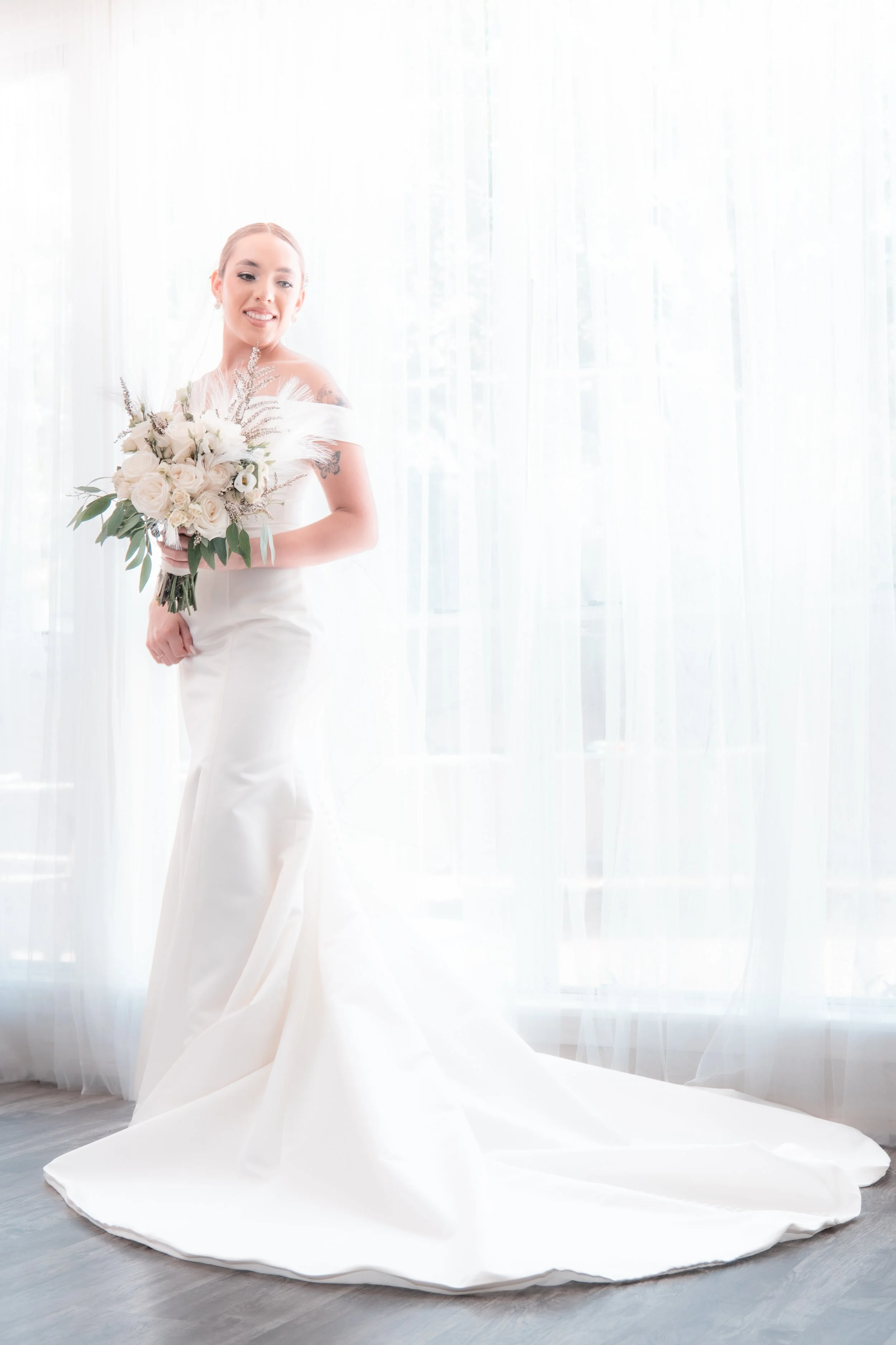 A bride in a white wedding gown holding a bouquet of flowers, standing near a window with sheer curtains.