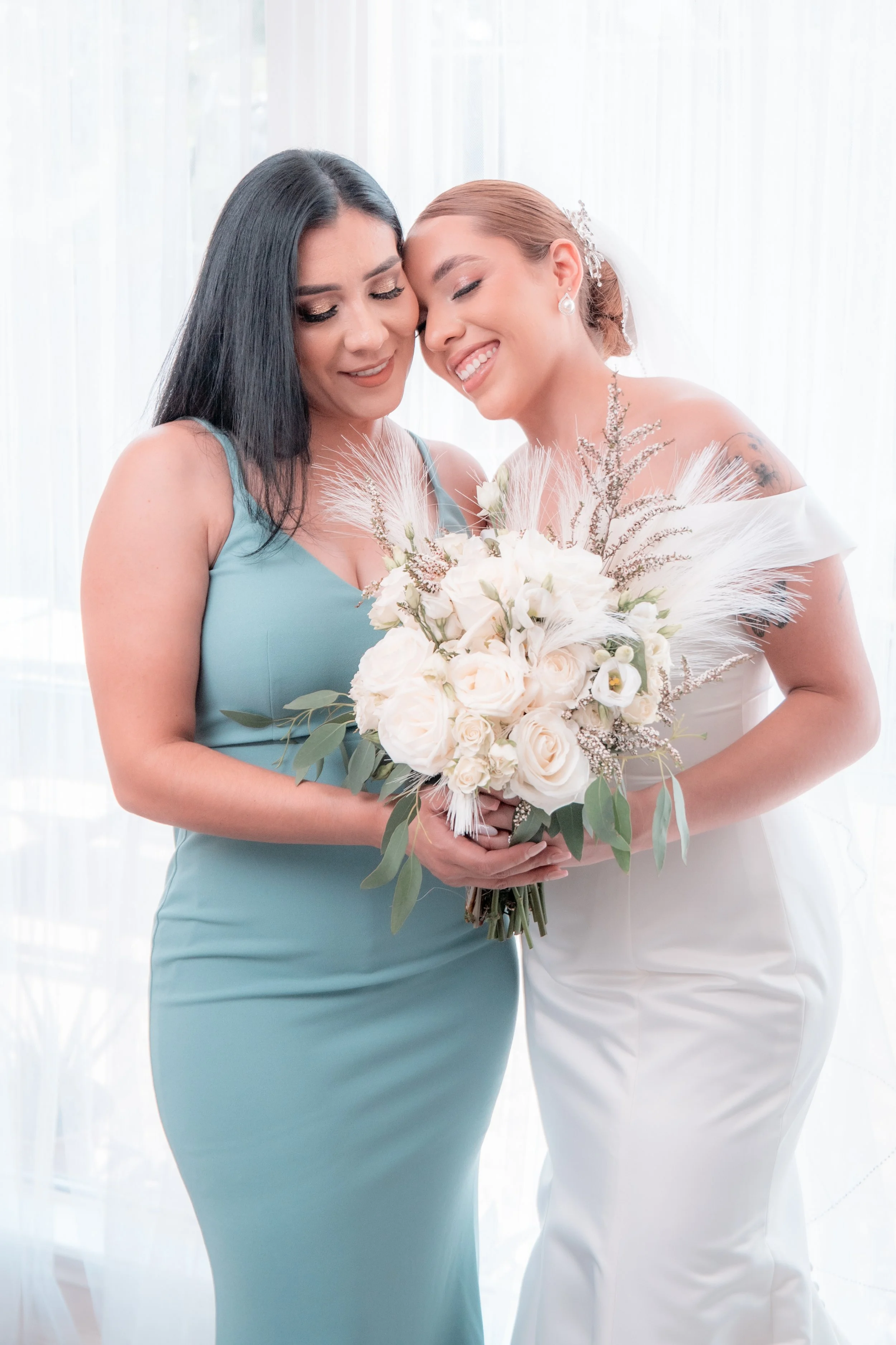 Two women, one in a wedding dress and the other in a light blue dress, share a tender moment while holding a bouquet of white and cream flowers, smiling with their foreheads touching.