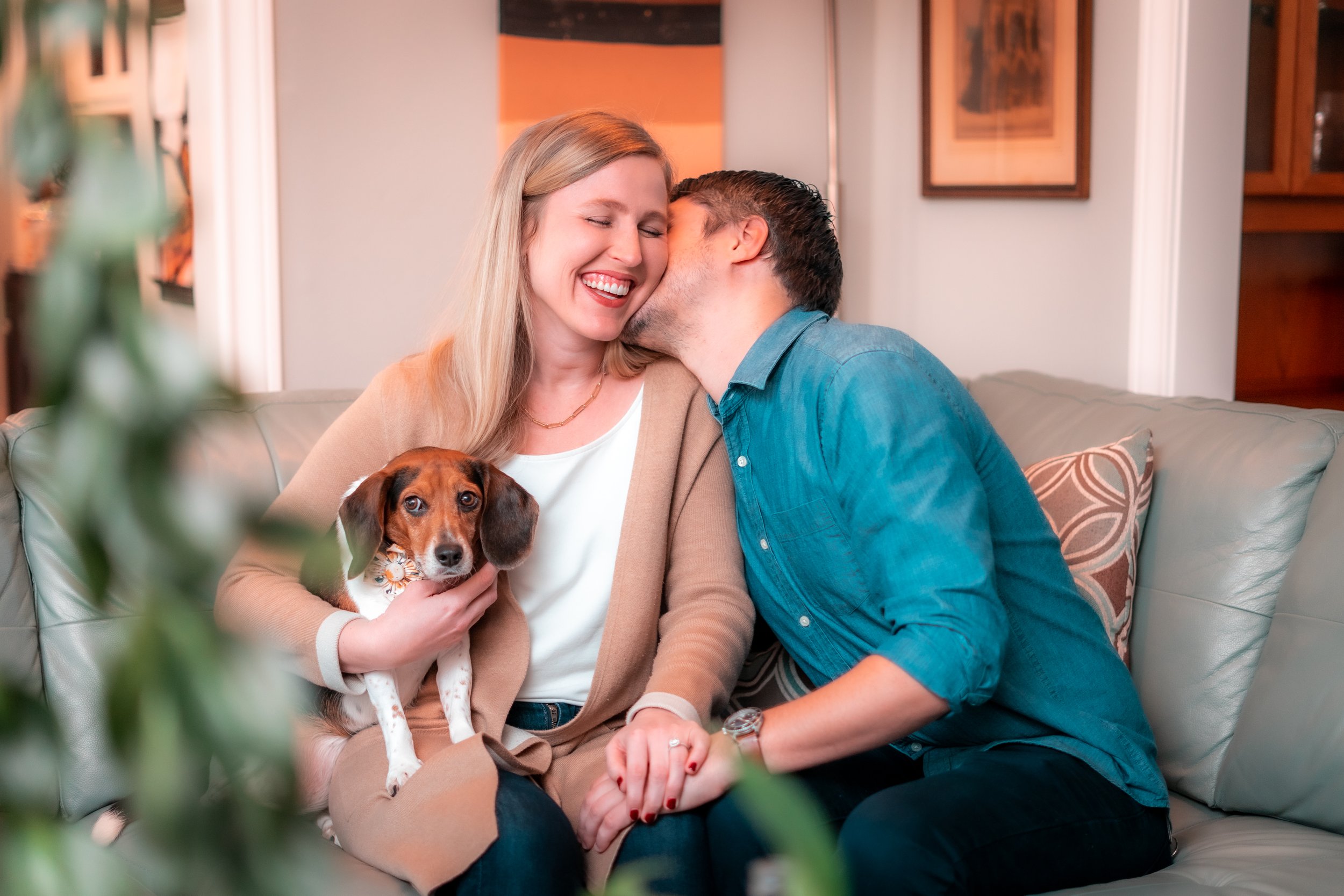 A happy couple sitting on a couch with their dog, sharing a kiss, in a cozy living room.