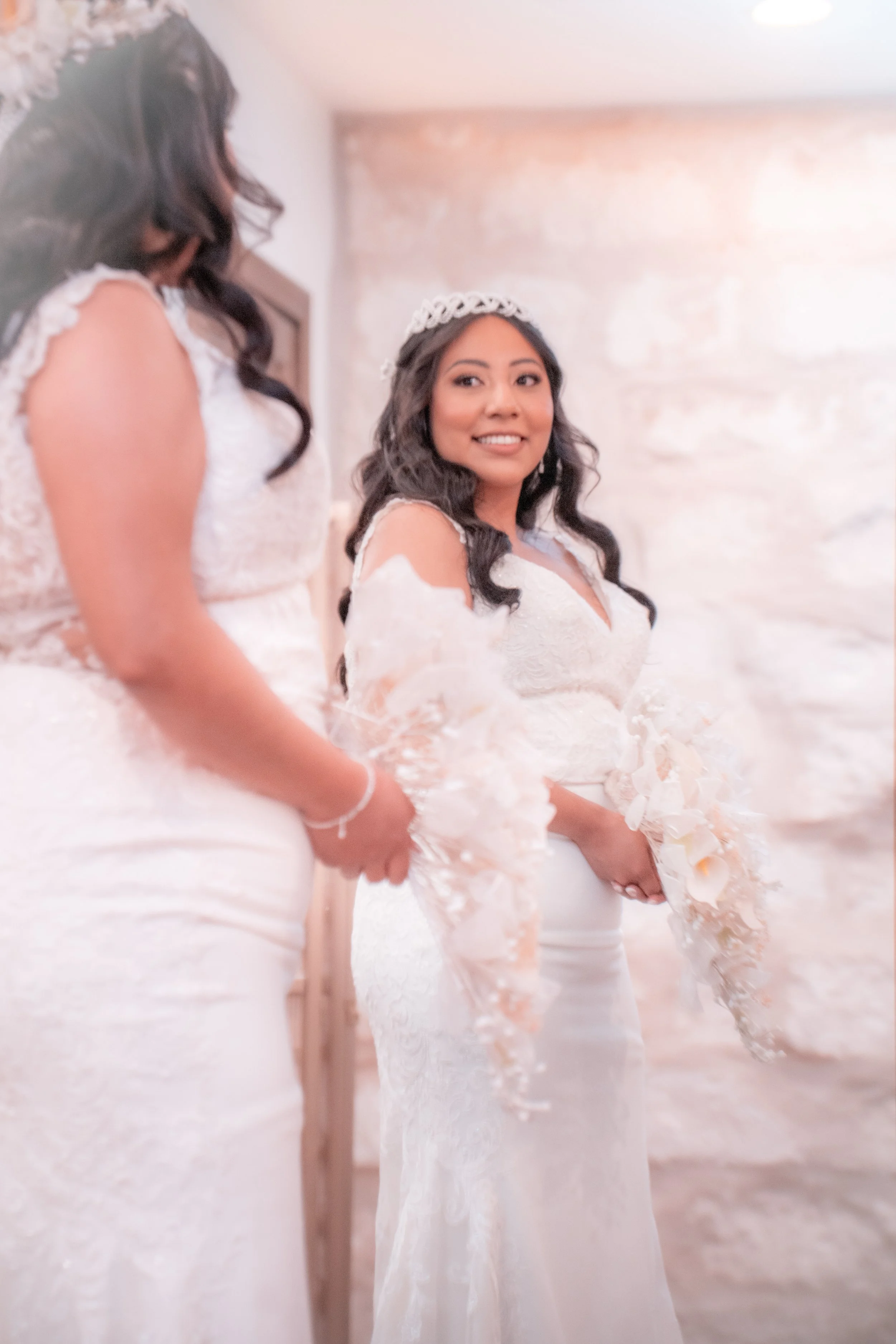 A bride holding a bouquet, smiling, wearing a lace wedding dress and a tiara, standing next to another woman also in a wedding dress, in a softly lit indoor setting with a stone wall background.