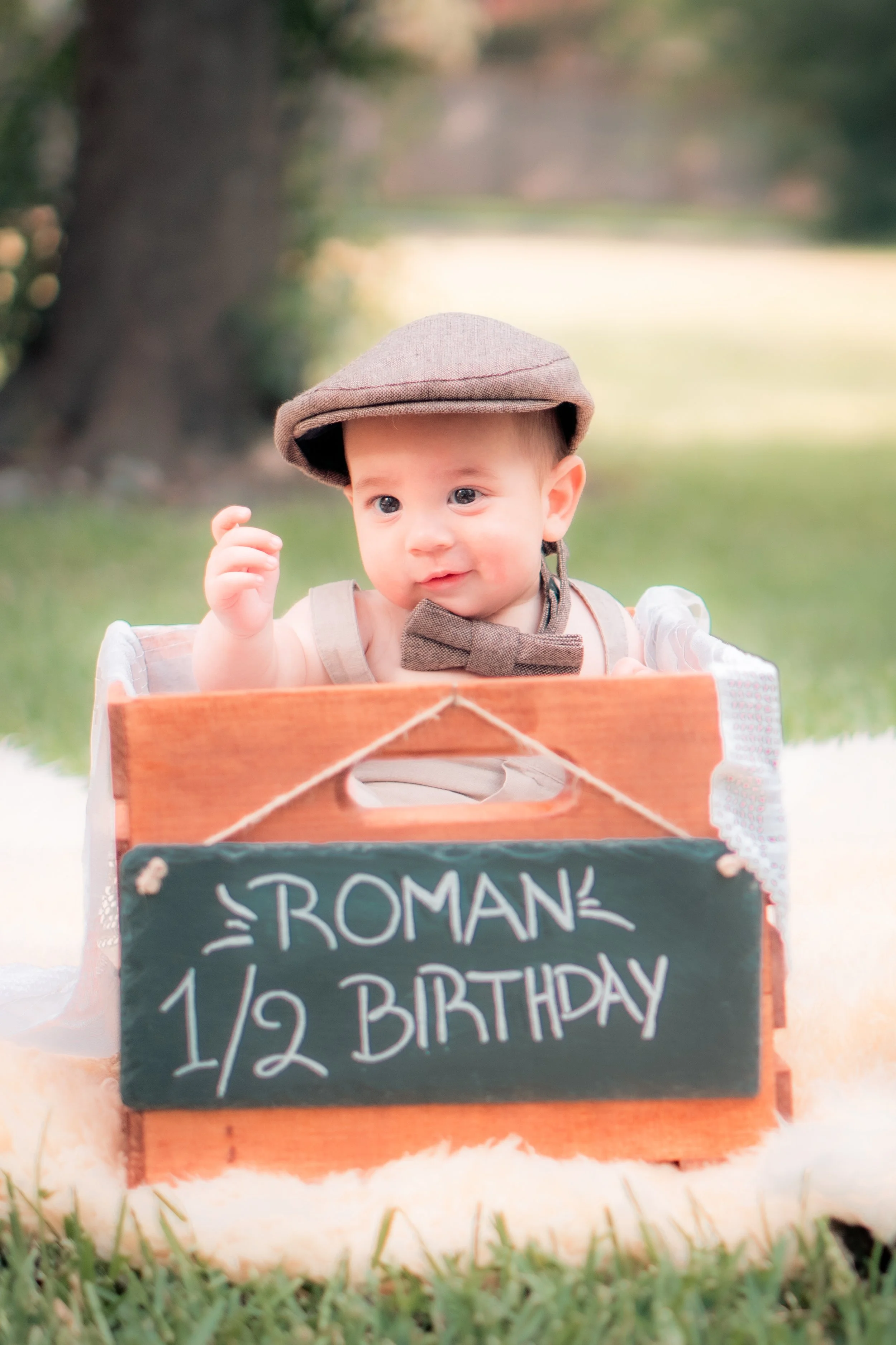 A baby sitting in a wooden box outdoors, wearing a brown cap and bow tie, with a sign that reads 'Roman's 1/2 Birthday' in front of him.