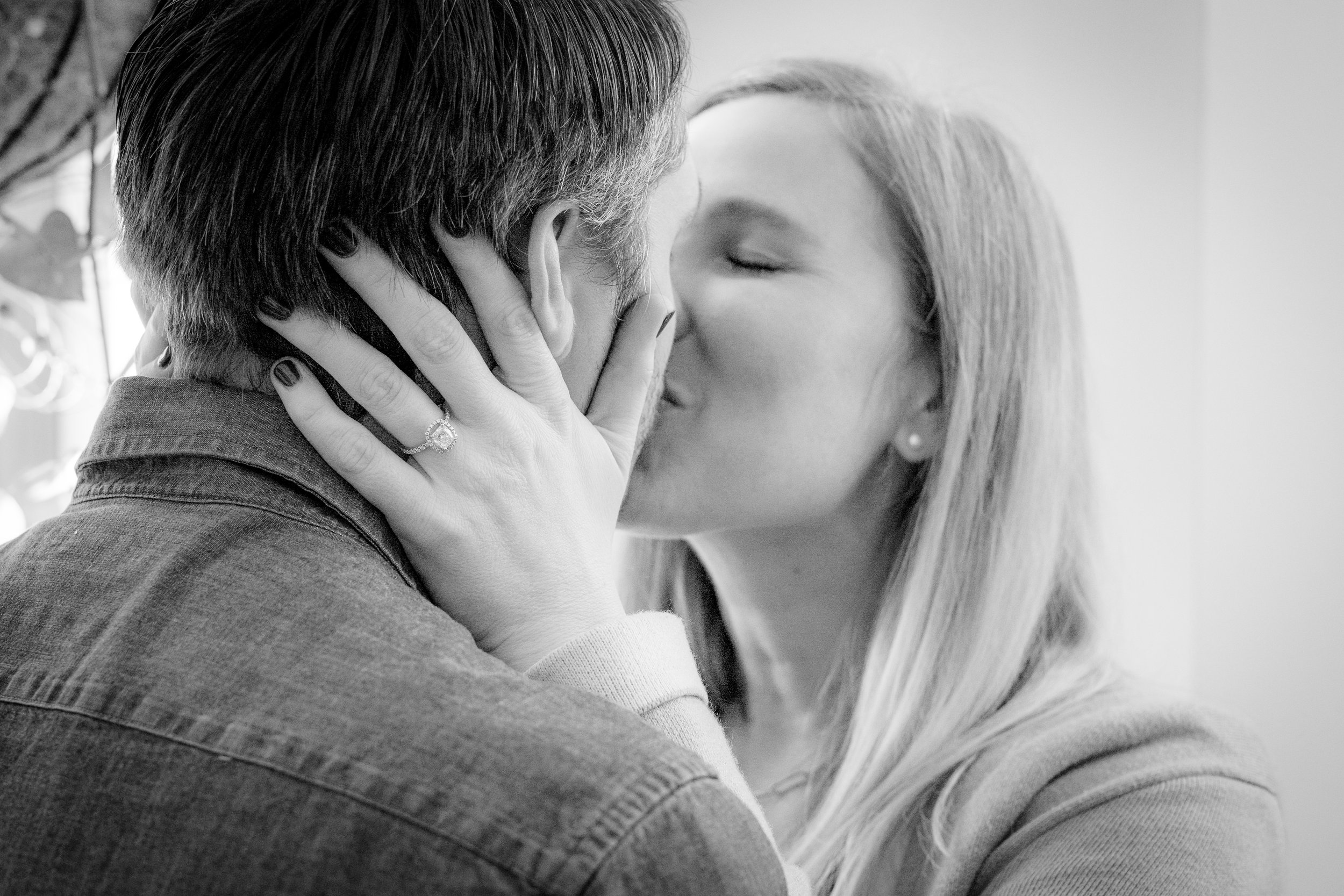 A woman is kissing a man on the lips, holding his face with both hands. She has long blonde hair, closed eyes, and is wearing a ring on her left hand, earrings, and a sweater. The man has short hair, is wearing a denim shirt, and his face is partiall