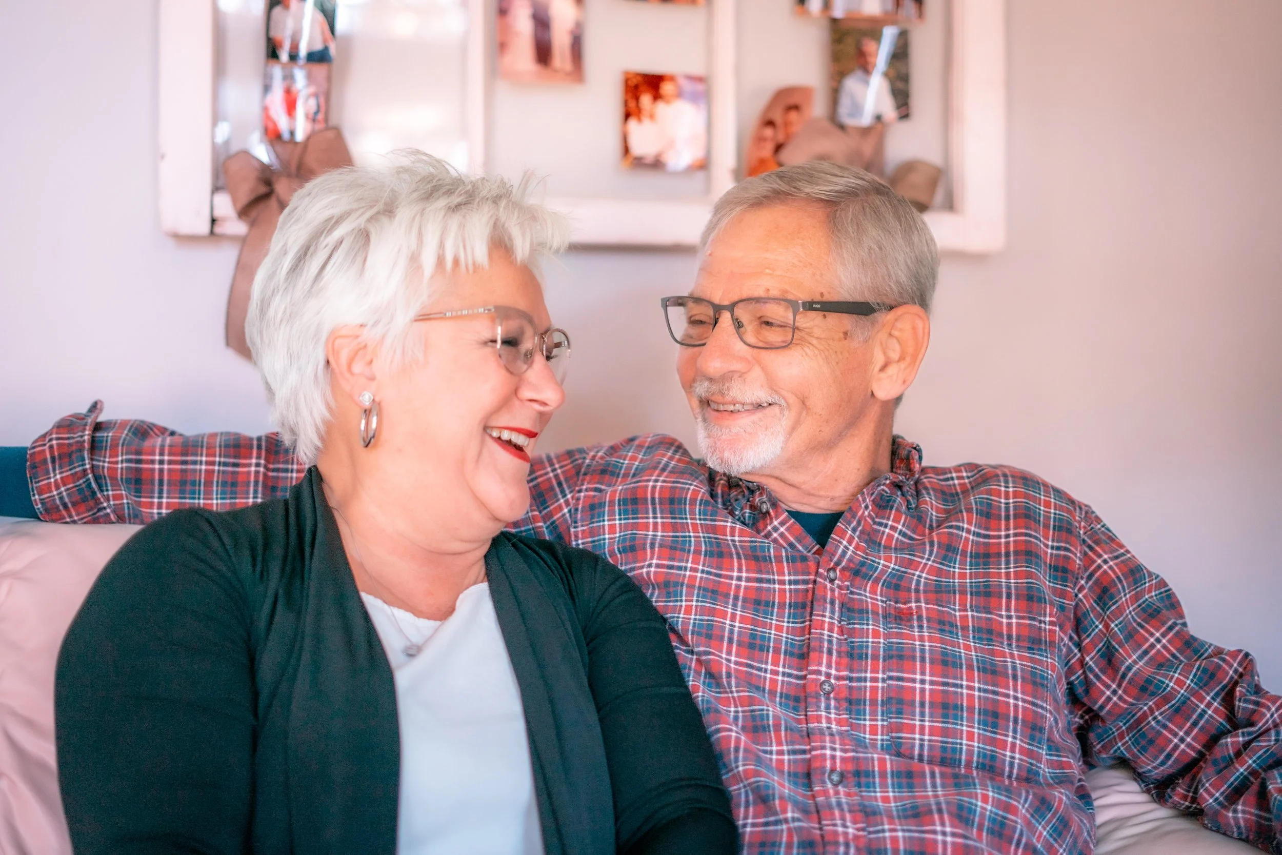 An elderly couple sitting close together on a couch, smiling and sharing a happy moment. The woman has short white hair, glasses, and is wearing a black jacket over a white top. The man has short gray hair, glasses, and is wearing a red plaid shirt. 