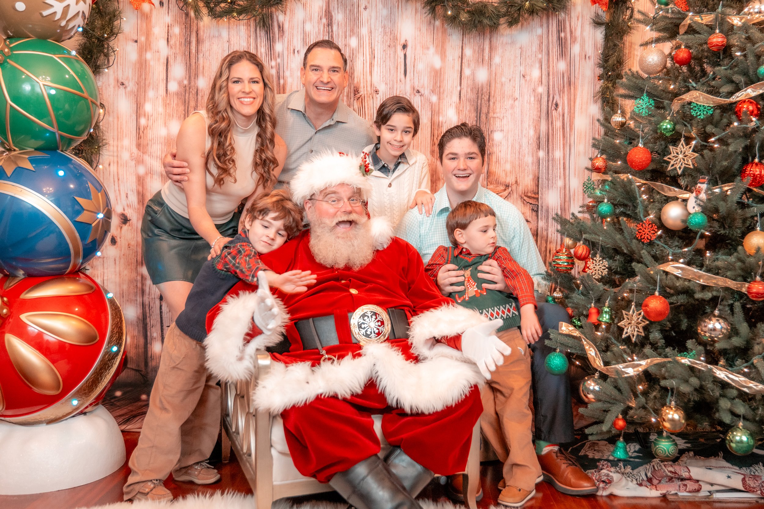 A family of seven posing with Santa Claus in front of a decorated Christmas tree and festive background.