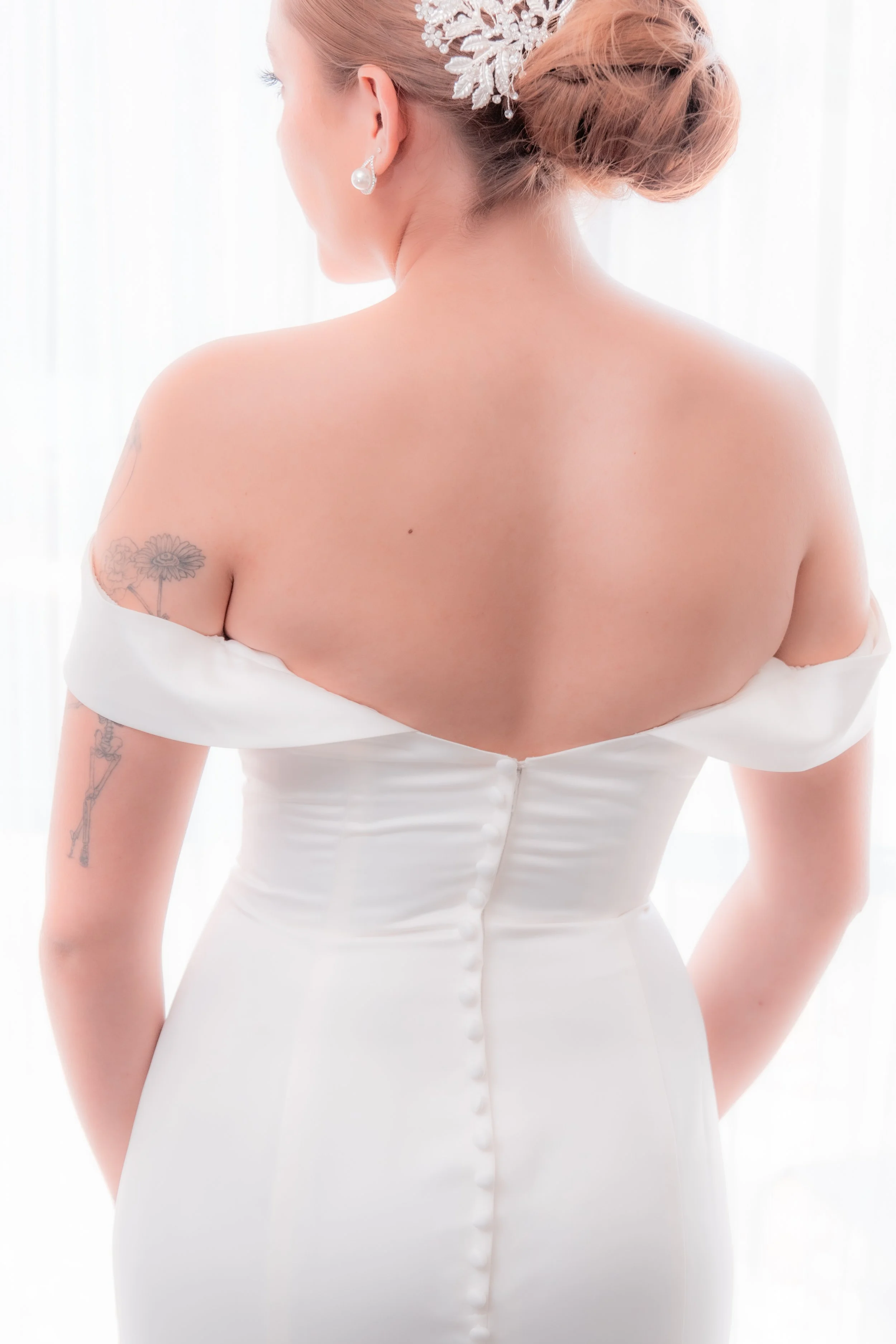 Back view of a woman in a white wedding dress, with styled hair, earrings, and a floral hair accessory, standing in front of a bright background.