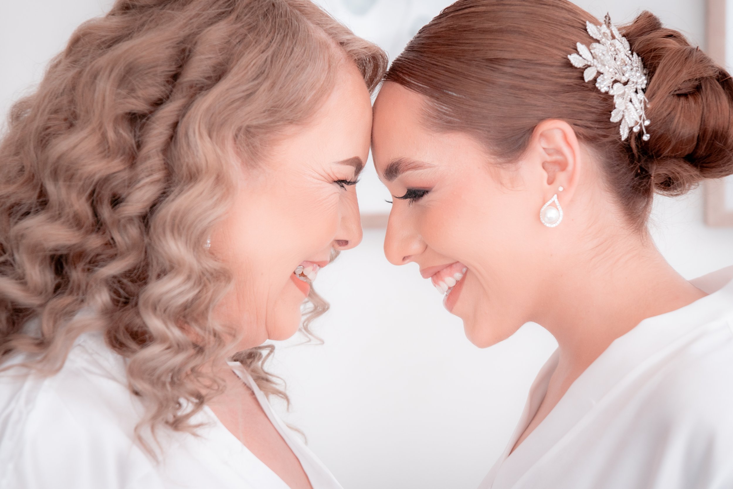 Two women with their foreheads and noses touching, smiling, dressed in white, with one wearing a wedding veil and jewelry, hinting at a wedding or special occasion.