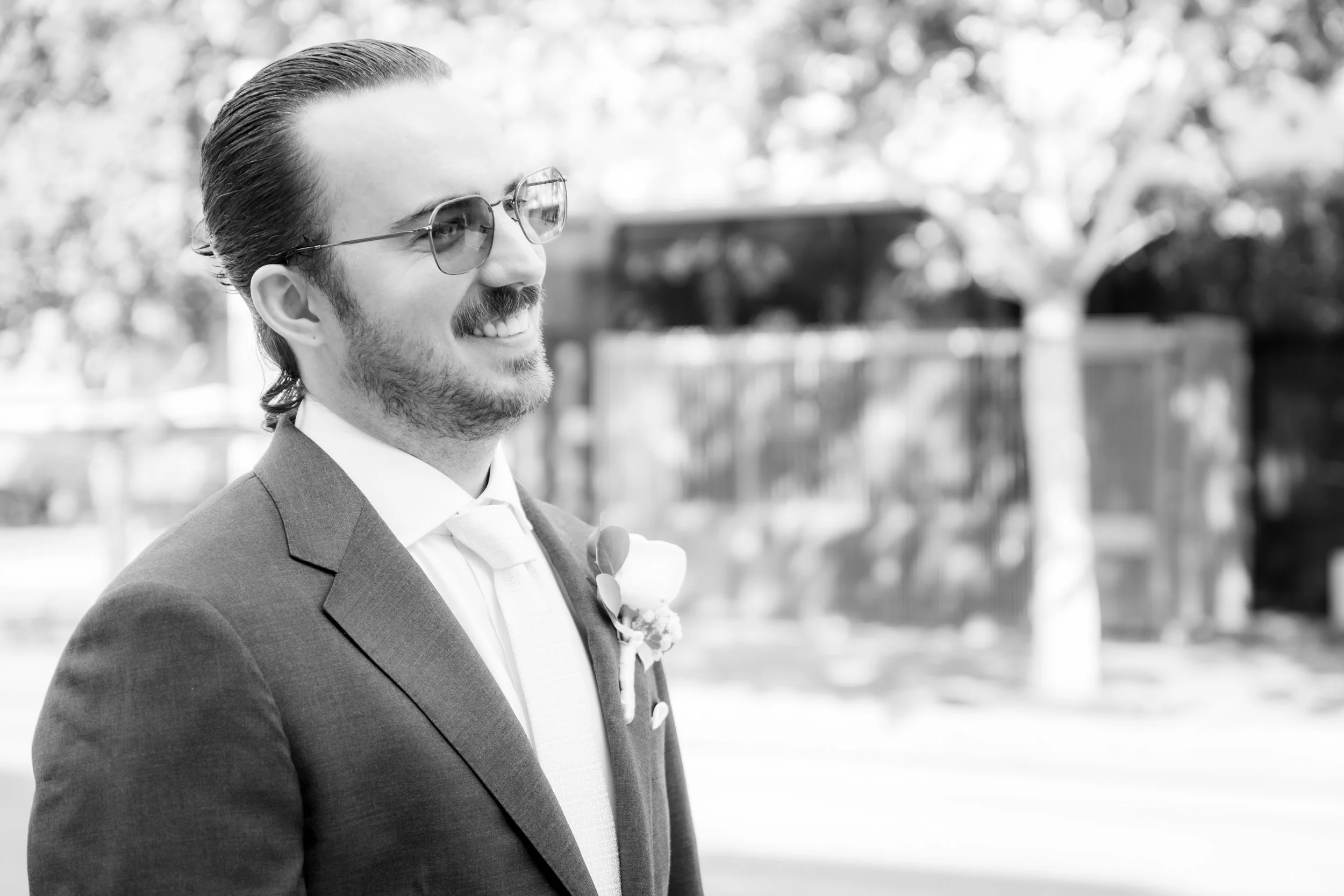 A man in a suit, wearing sunglasses and a boutonniere, smiling outdoors.