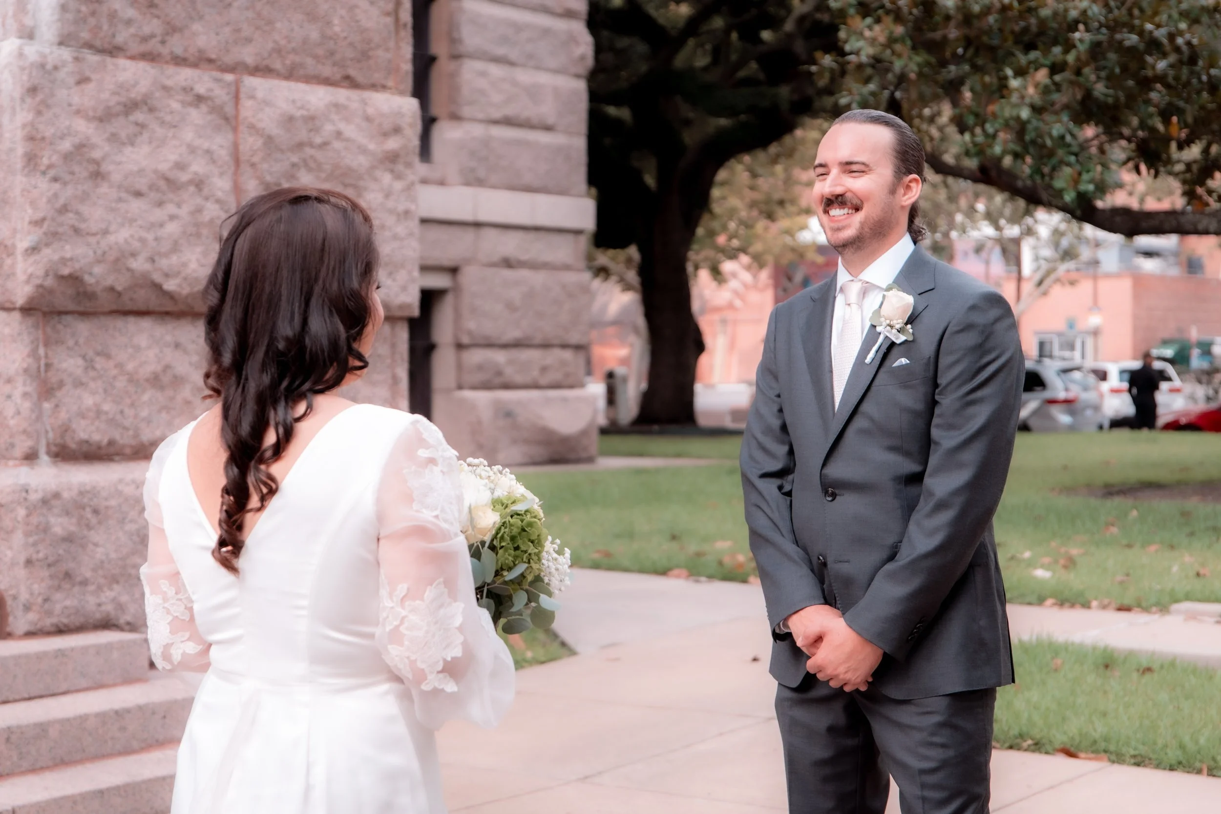 A groom in a gray suit and a bride in a white dress holding a bouquet, standing outdoors in front of a stone building, sharing a moment at their wedding.