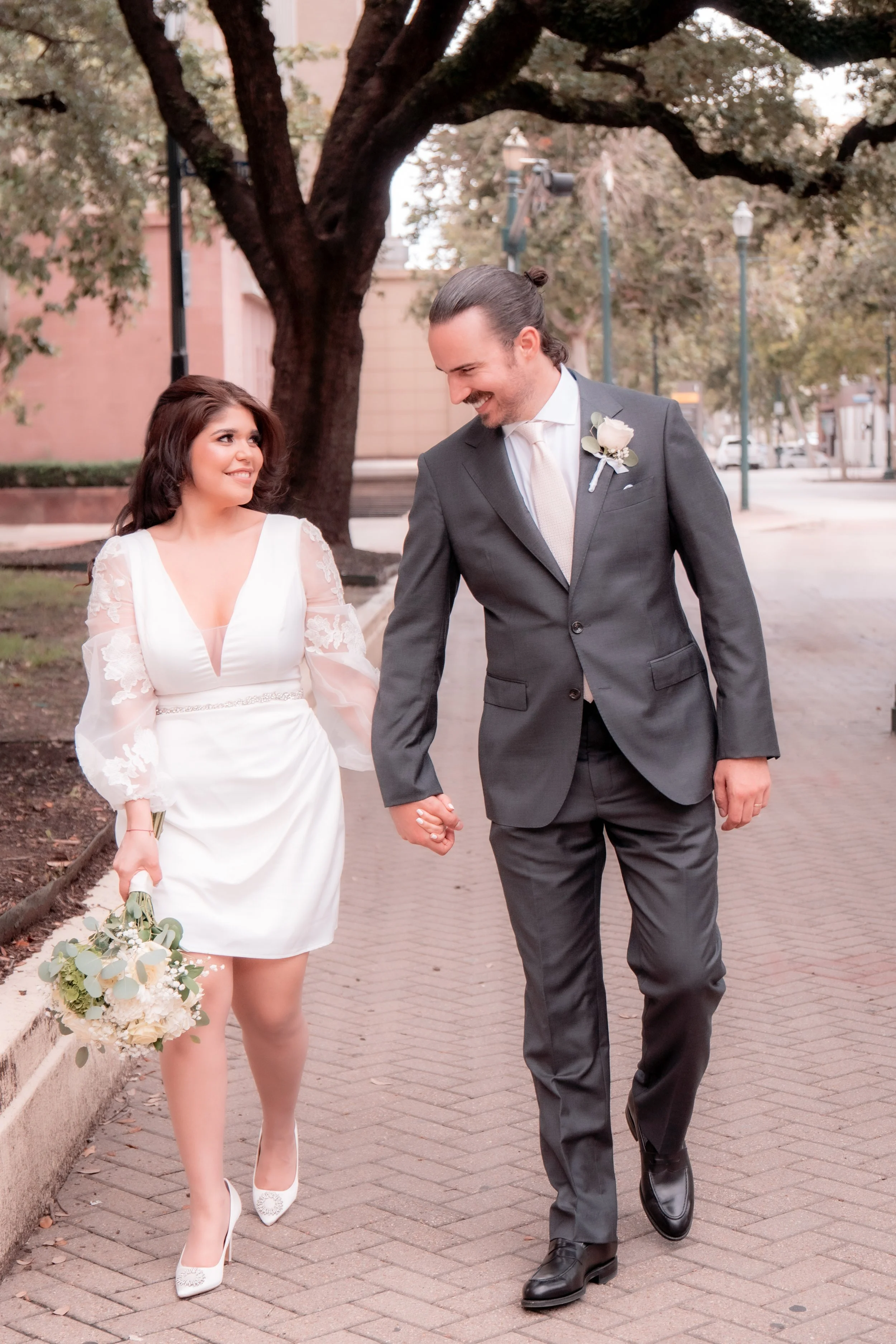 A bride and groom walking hand in hand on a city street, smiling and looking at each other. The bride wears a white dress with lace sleeves and holds a bouquet. The groom wears a gray suit with a white shirt and tie.