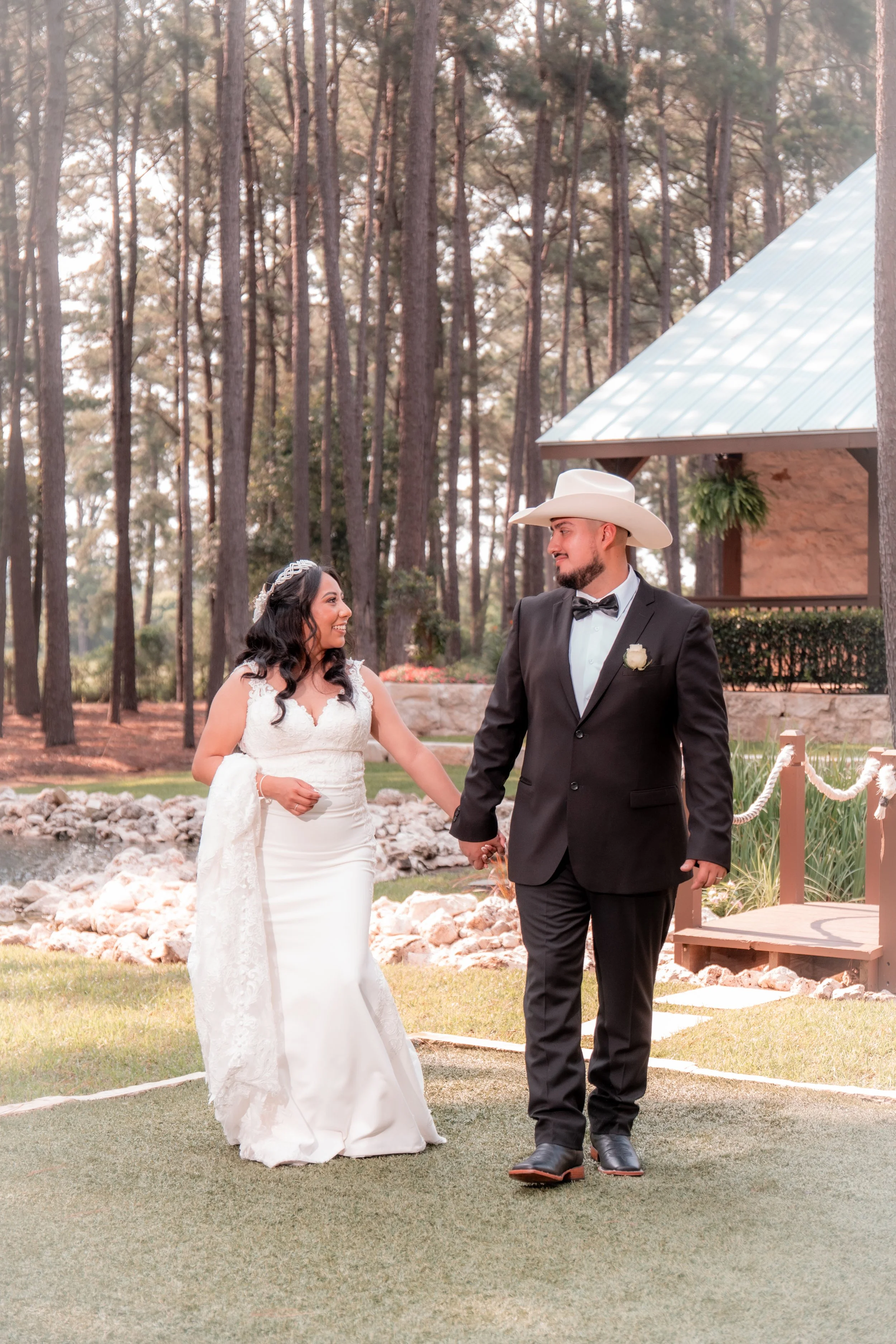 A bride and groom walking hand in hand outdoors during their wedding ceremony in a wooded area, with the bride in a white dress and the groom in a black suit and cowboy hat.