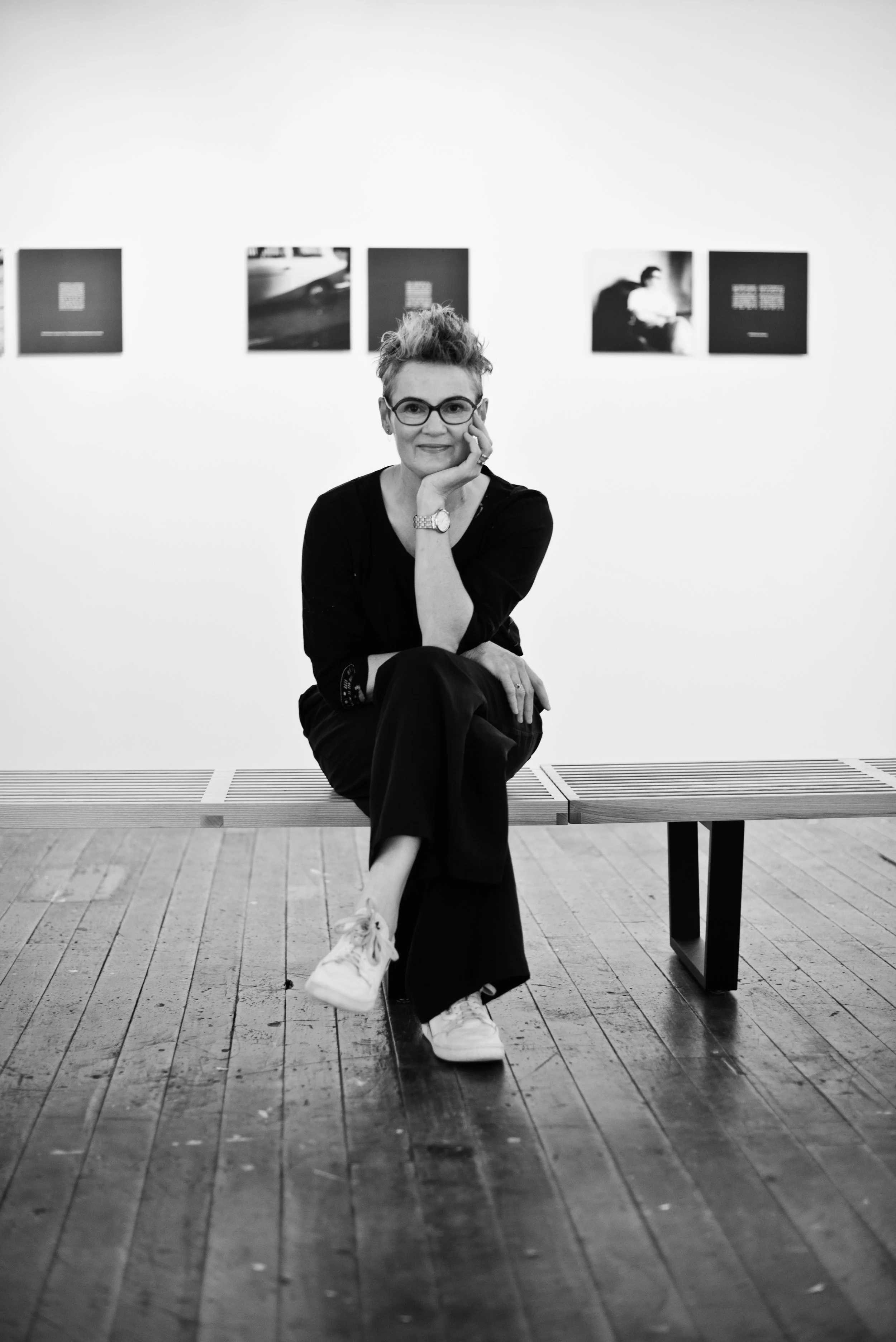 Catherine Russ seated on a bench in a gallery, chin resting on her hand, framed black-and-white works on the wall behind her.