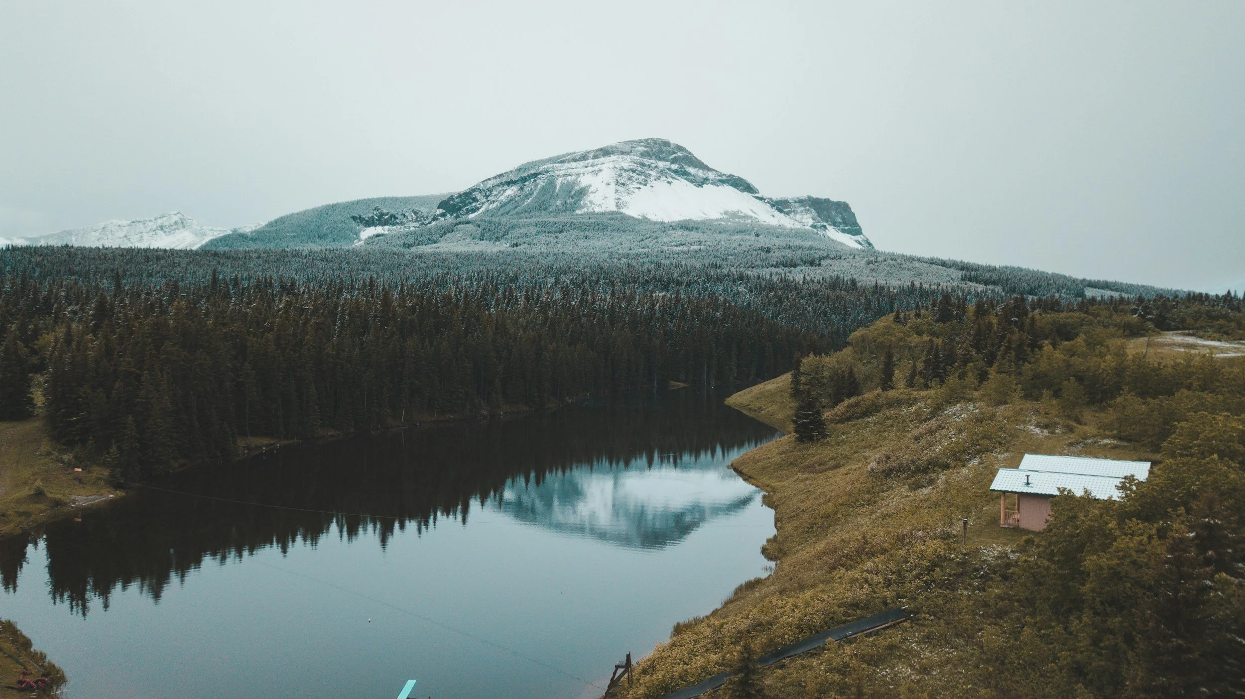 A mountain with snow on its peak and slopes, surrounded by a forest of coniferous trees, with a lake reflecting the mountain and trees in the foreground. A small house and some rooftops are visible near the bottom right corner.