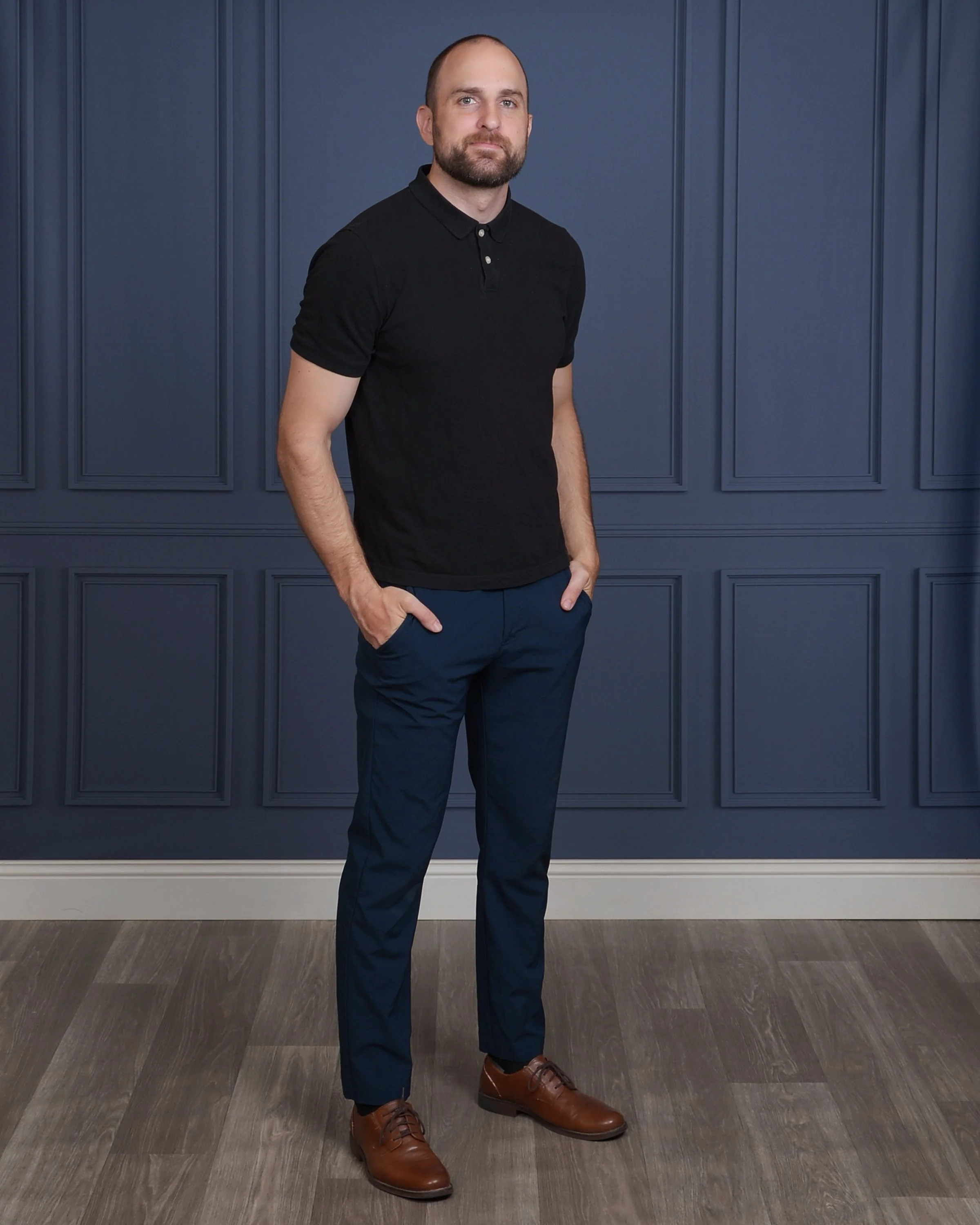A man with a beard, dressed in a black polo shirt and navy blue pants, standing indoors against a dark blue paneled wall with his left hand in his pocket and looking at the camera.