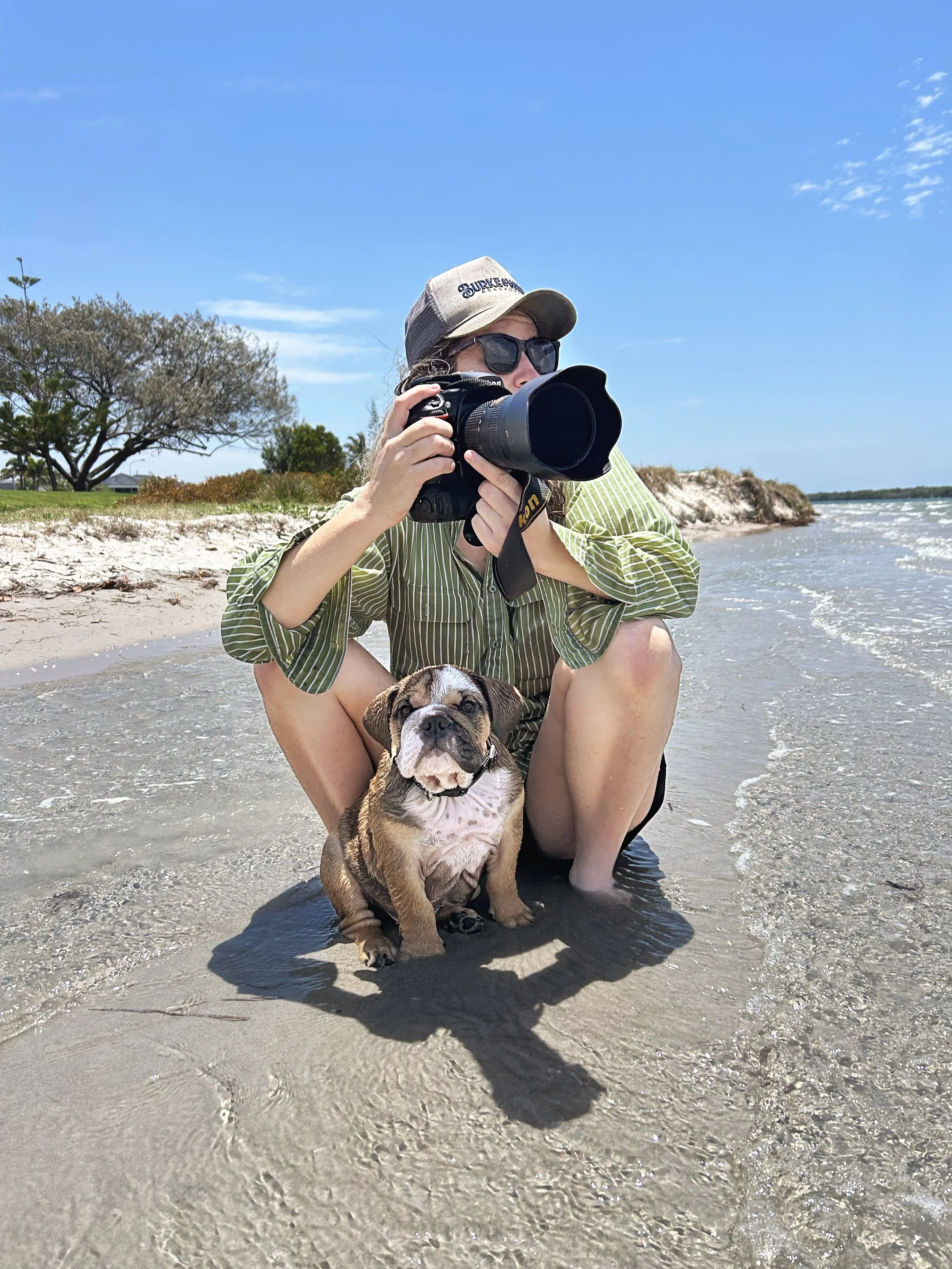 Person in green striped shirt, sunglasses, and a cap sitting in shallow water at the beach, taking photos with a large camera, with a smiling puppy sitting in front of them.