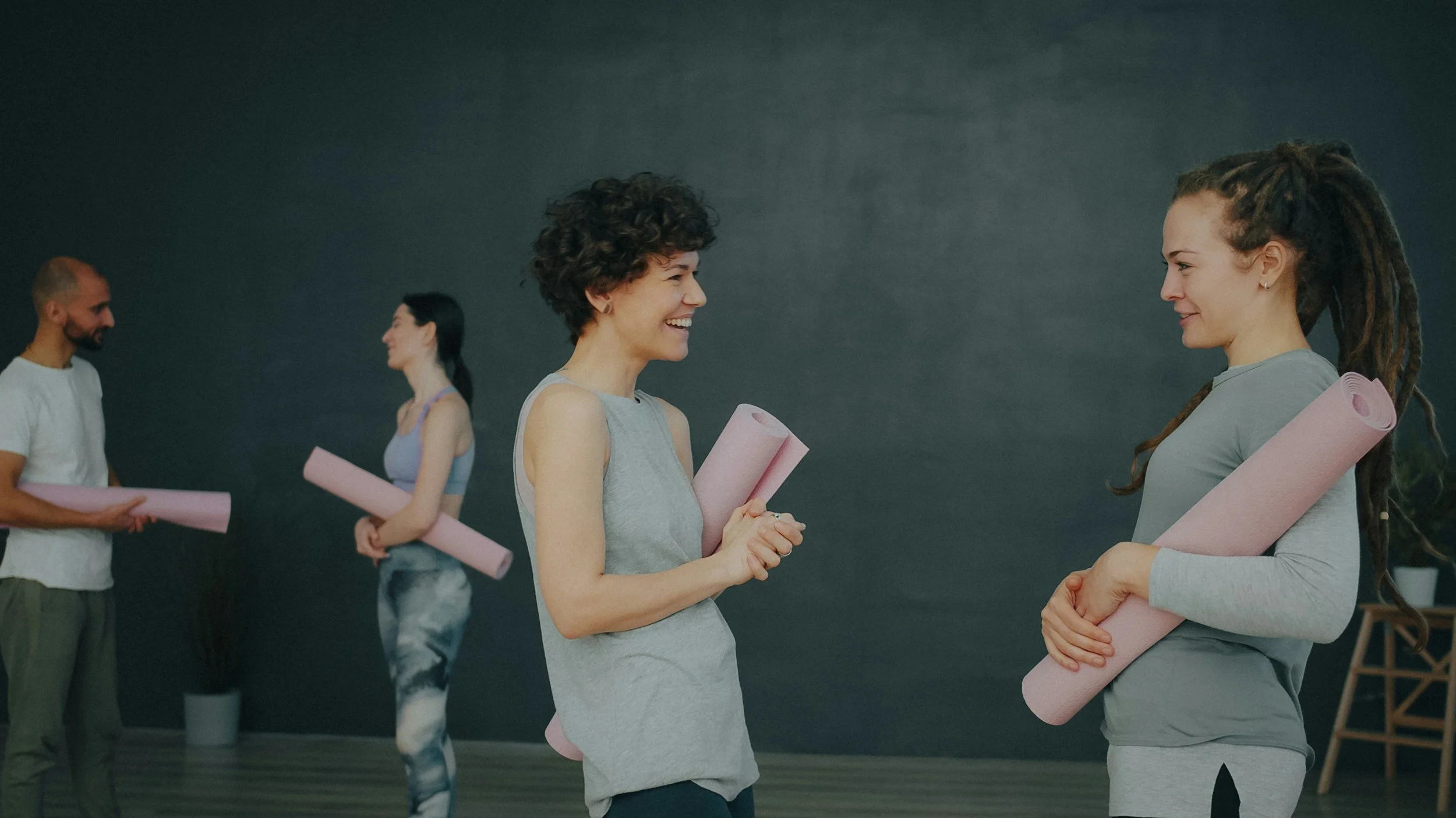 Two women with yoga mats talking and smiling while a group in the background holds pink yoga mats.