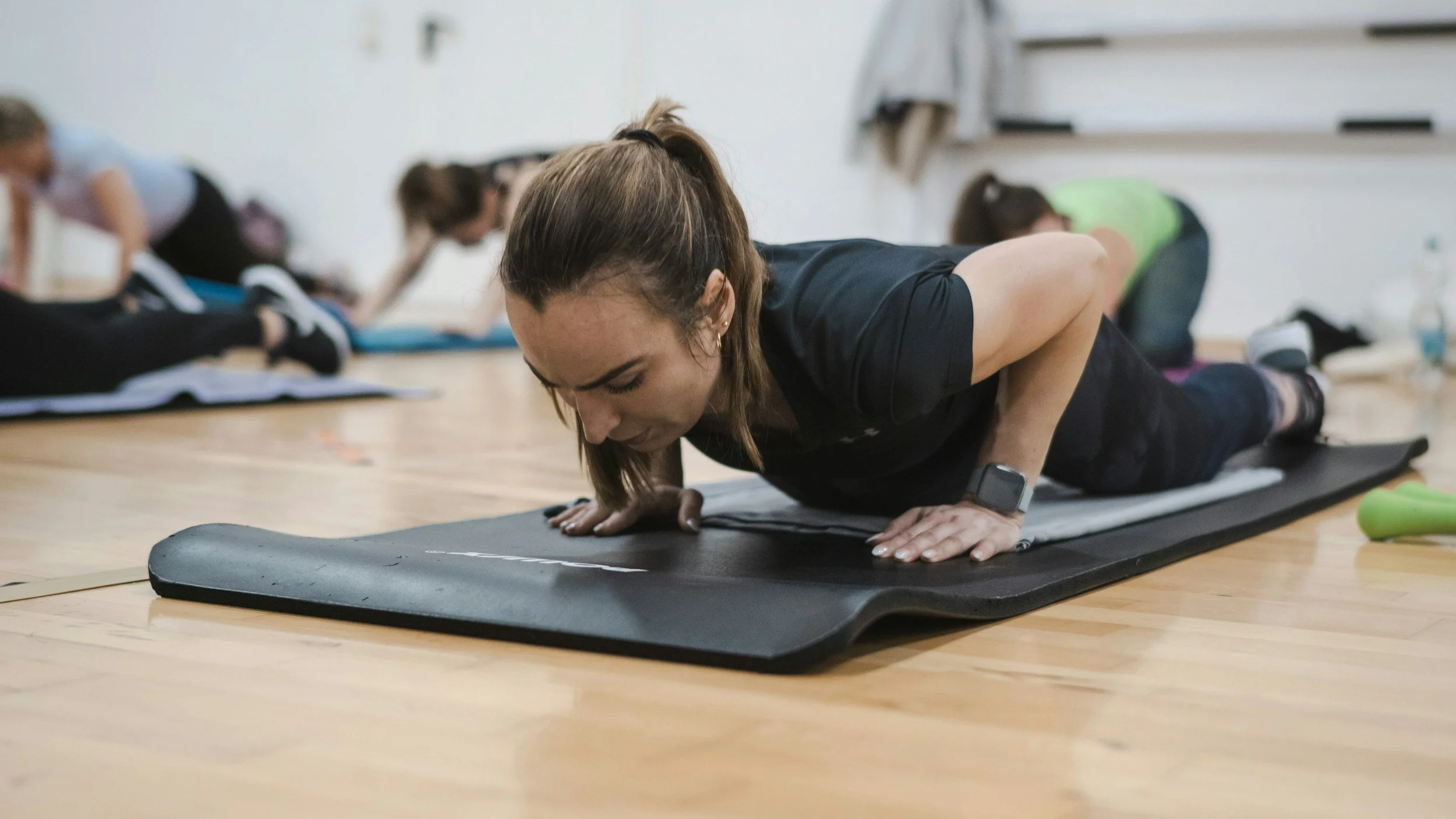 Woman doing a plank exercise on a black yoga mat in a fitness class, with other participants in the background