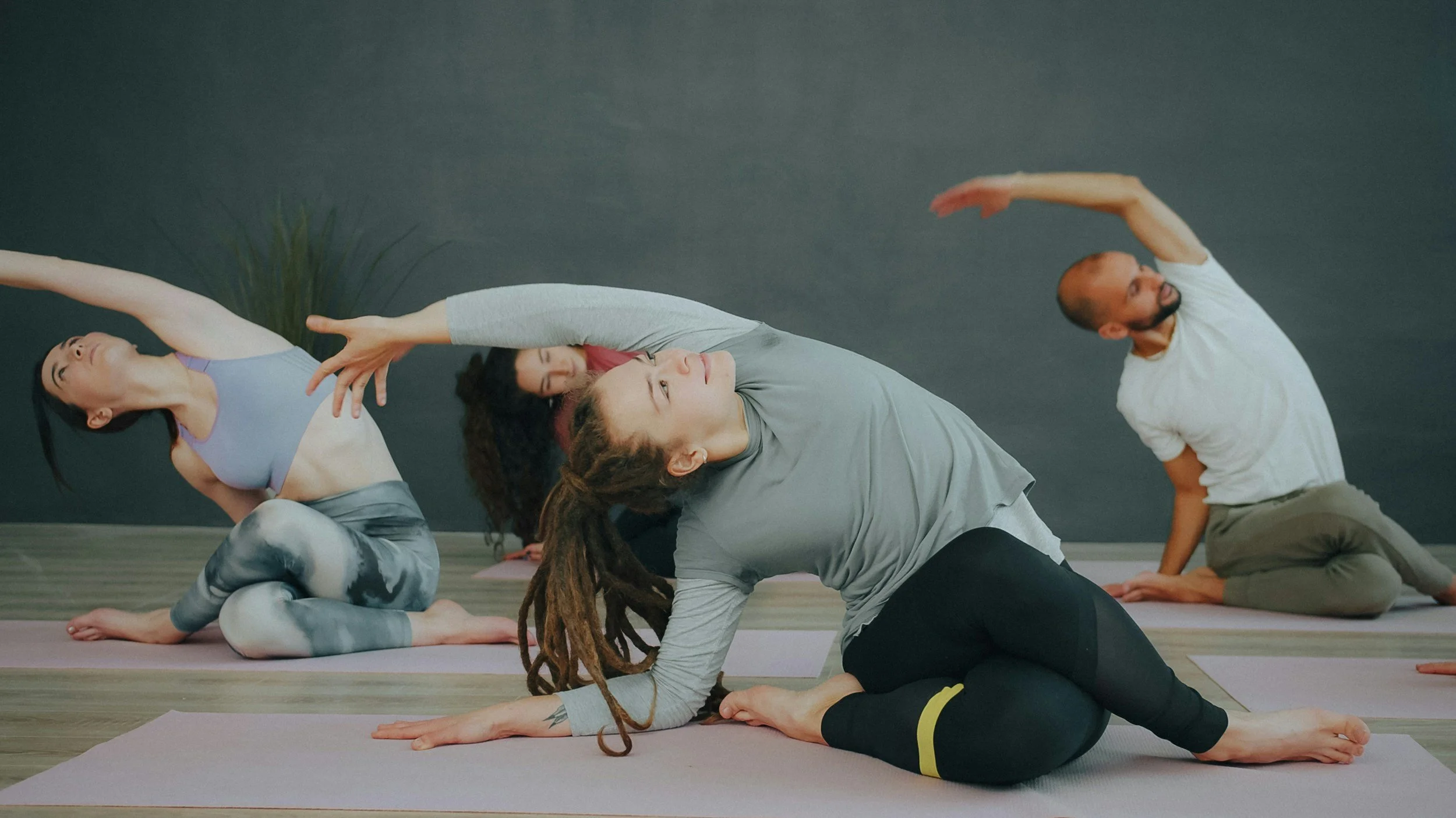Group of people practicing yoga in a studio, sitting on mats with one arm raised overhead, in a side stretch pose.