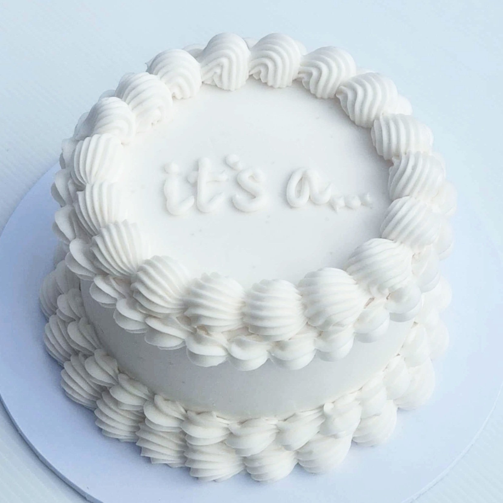 White cake with "it's a..." written on top, decorated with piped frosting.