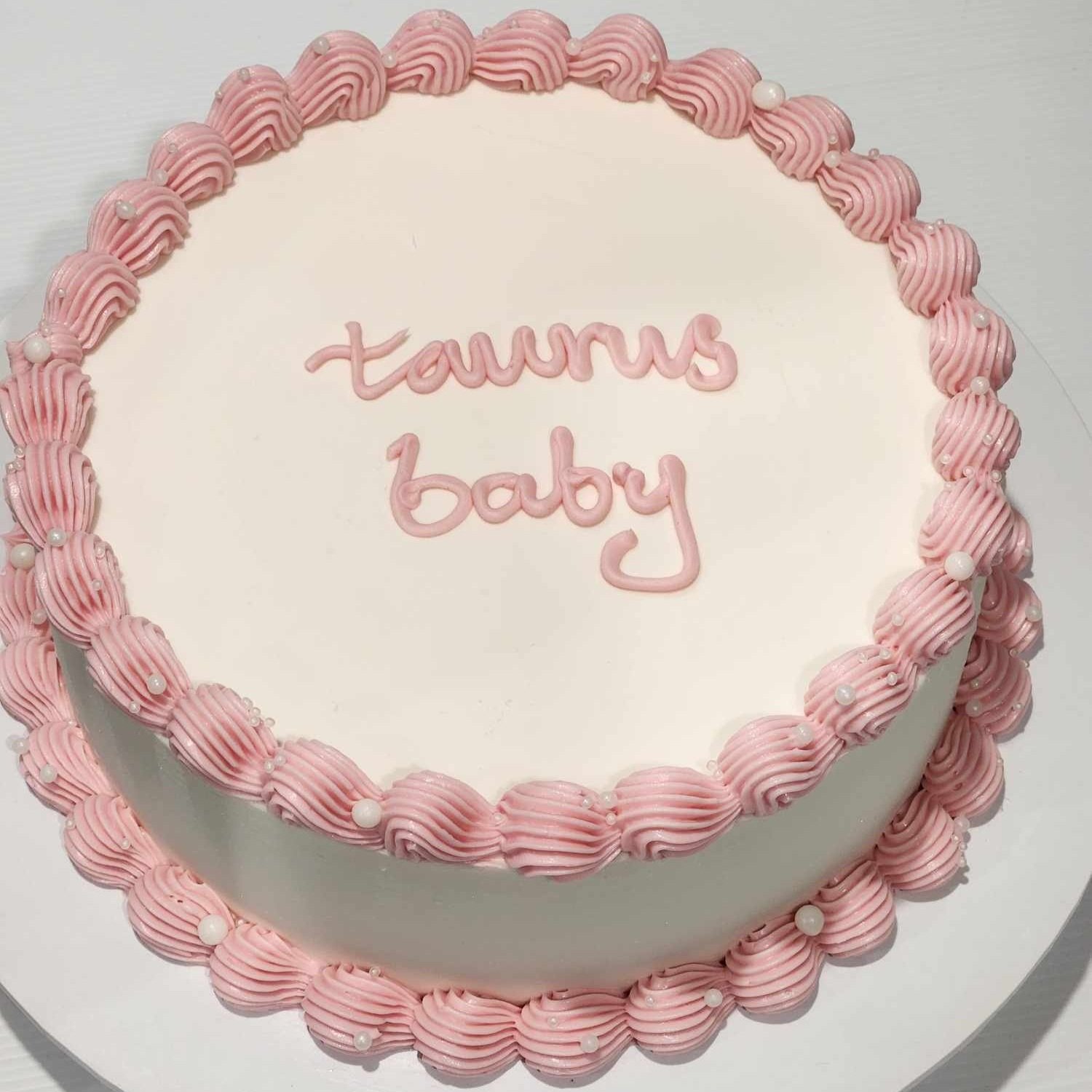 A round cake with pink frosting piped along the edges and the words "taurus baby" written on top.