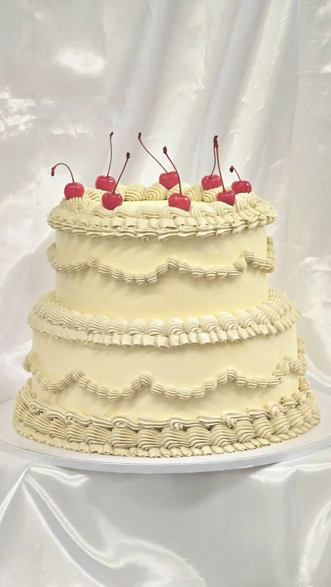 Three-tiered white cake decorated with piped cream, topped with cherries, on a white plate against a white satin background.