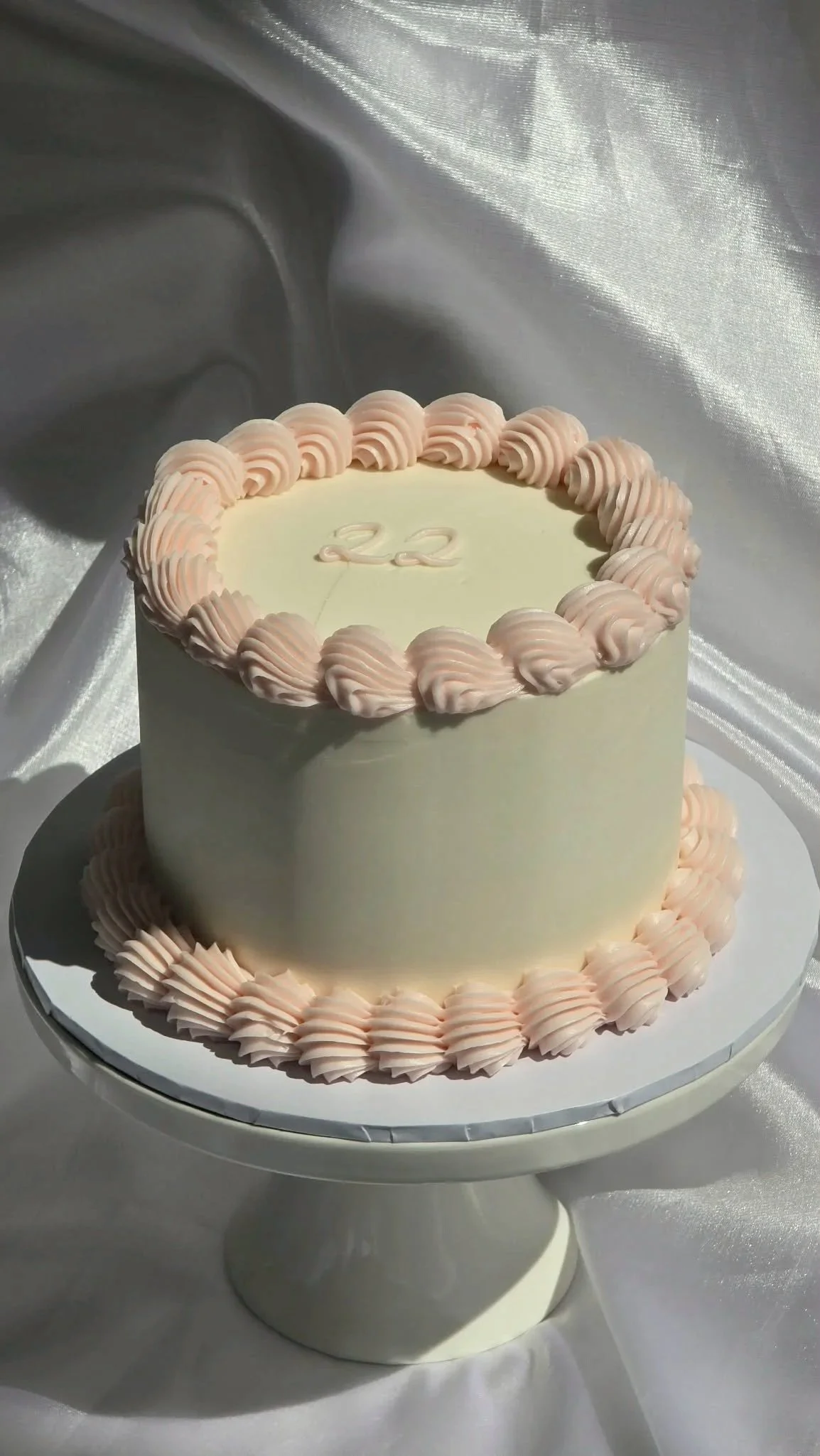 A two-tiered cake with a cream-colored base, decorated with pale pink piped rosettes around the edges, and the number 22 written on top in white. The cake is on a white cake stand with a satin-like fabric in the background.