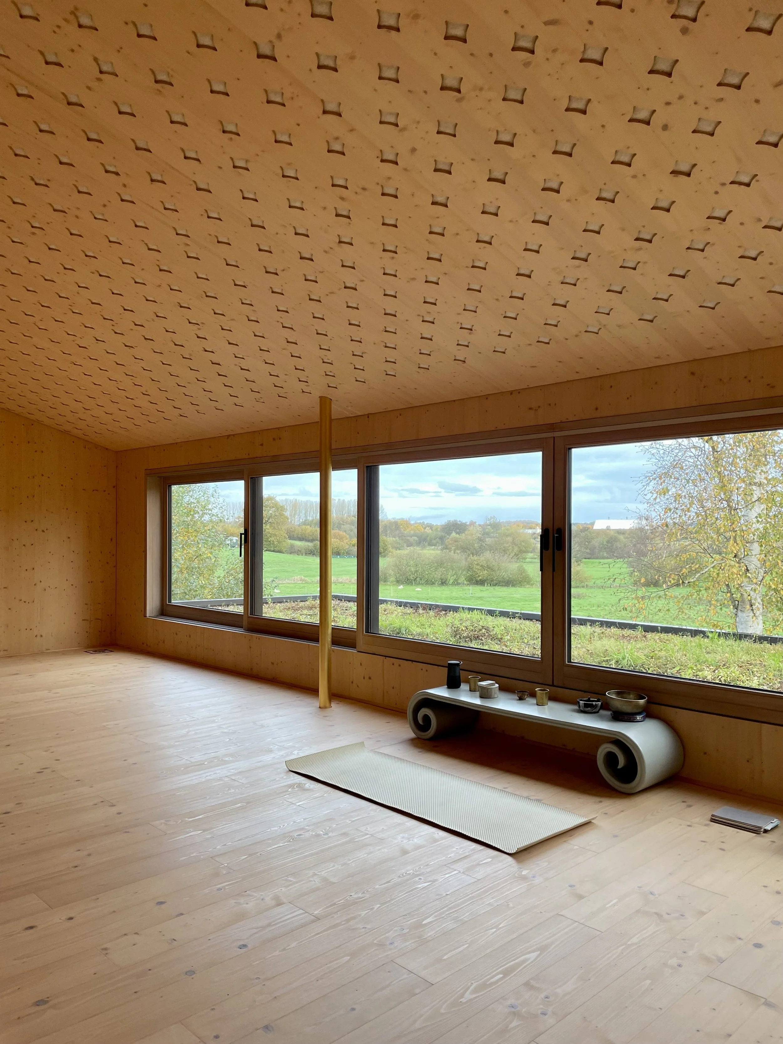 Innenraum eines Holzhauses mit großen Fenstern, Blick auf eine grüne Landschaft. Im Raum liegen noch keine Möbel. An einer Wand steht eine große Fensterbank mit Dekorationsgegenständen, darunter eine Spirale. Der Raum hat eine Holzboden- und Deckenverkleidung.