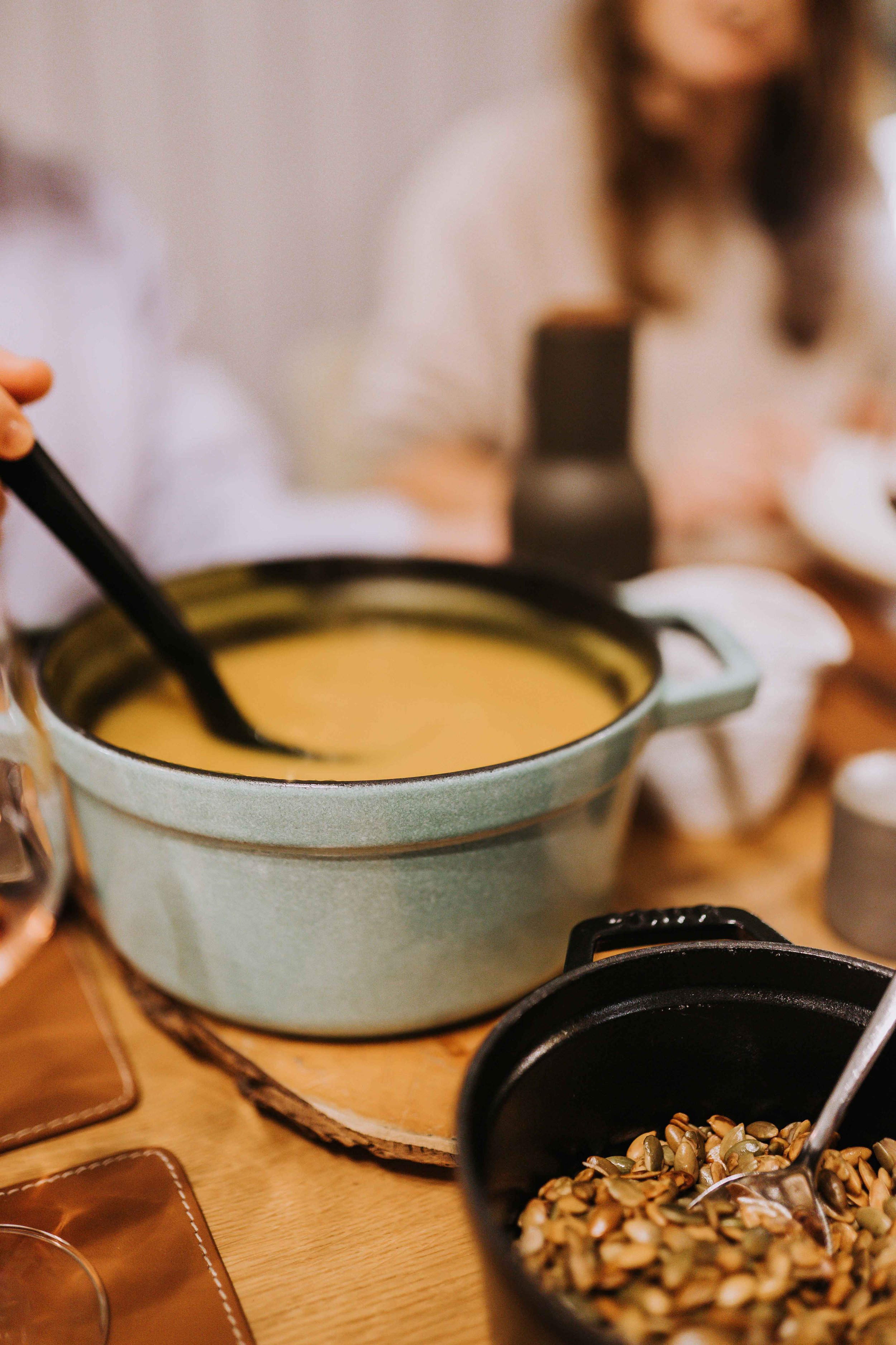 Eine Schüssel mit brauner Suppe, ein Becher mit Sonnenblumenkernen, Hefe, jemand in der Küche, Küchenutensilien auf einem Holztisch.