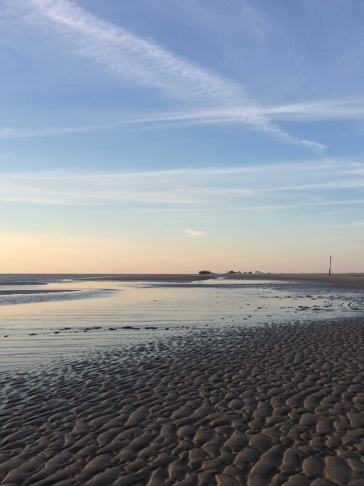Ein Strand bei Sonnenuntergang mit sandigem Ufer, ruhigem Wasser und einem offenen Himmel mit weißen Wolken.